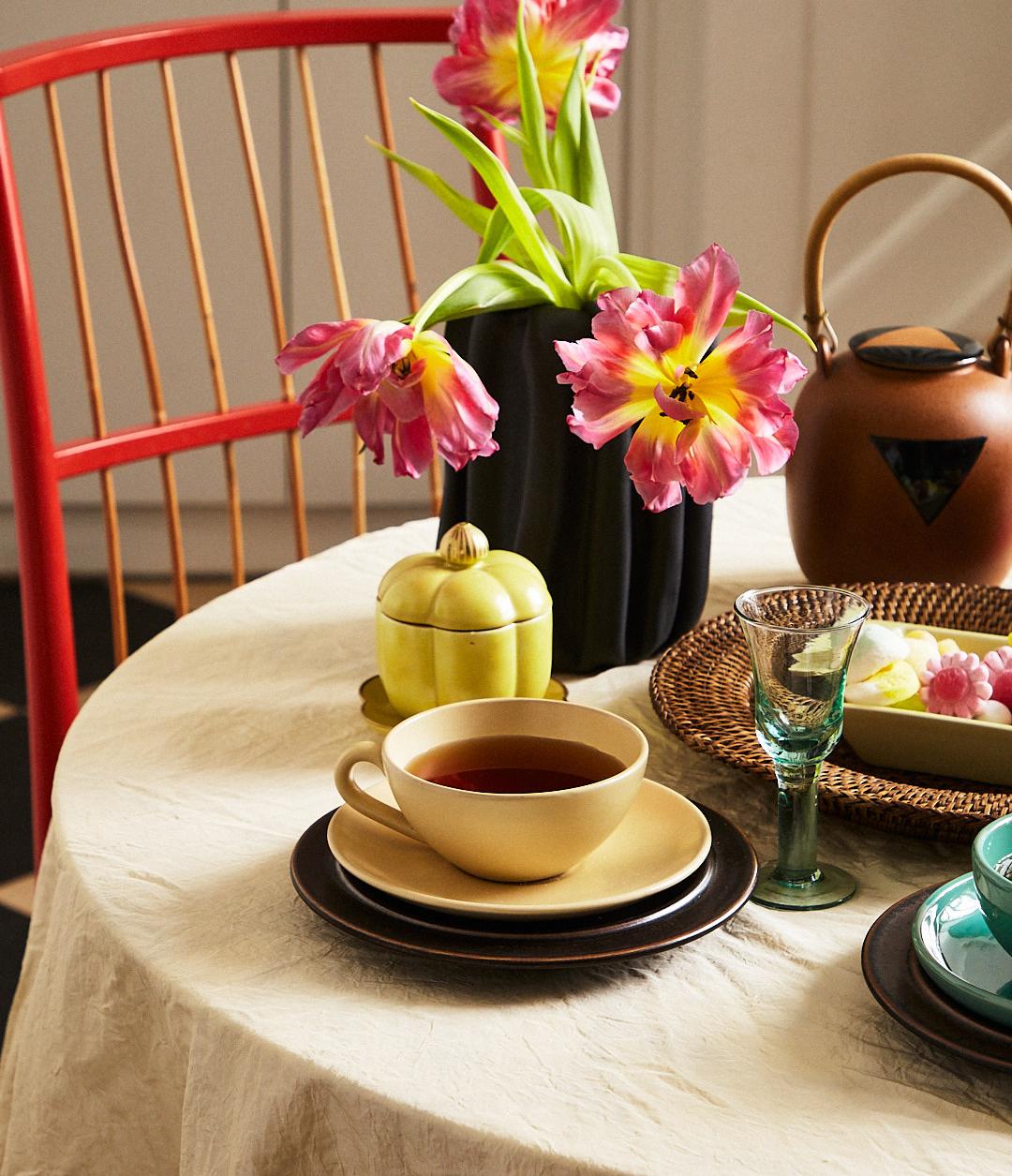 Ett bord dukat för te med en svart vas med rosa och gula tulpaner, en brun tekanna, en tekopp och färgglada karameller, bredvid en röd trästol
