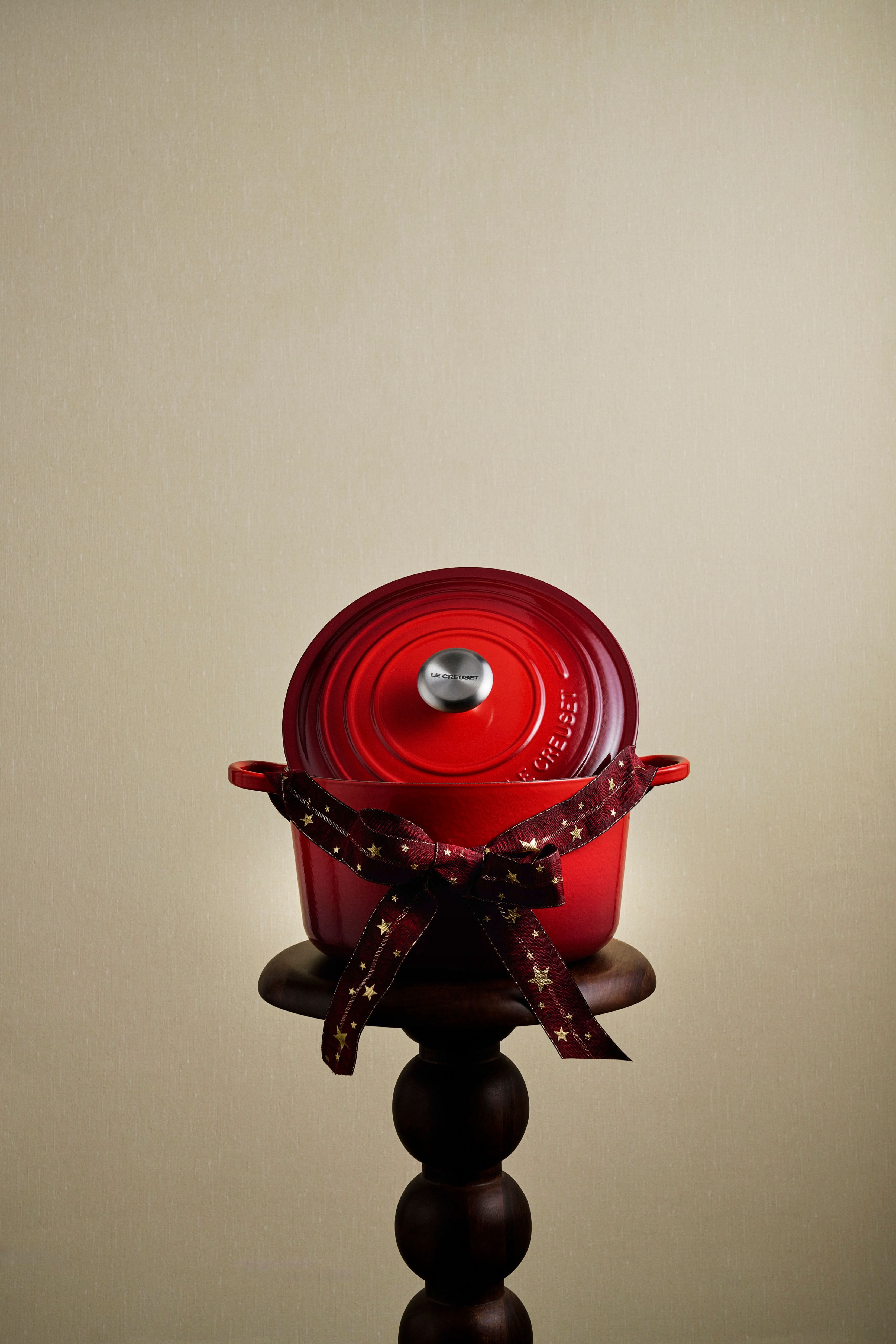 A red Le Creuset Dutch oven, tied with a star-patterned ribbon, sits on a wooden pedestal.