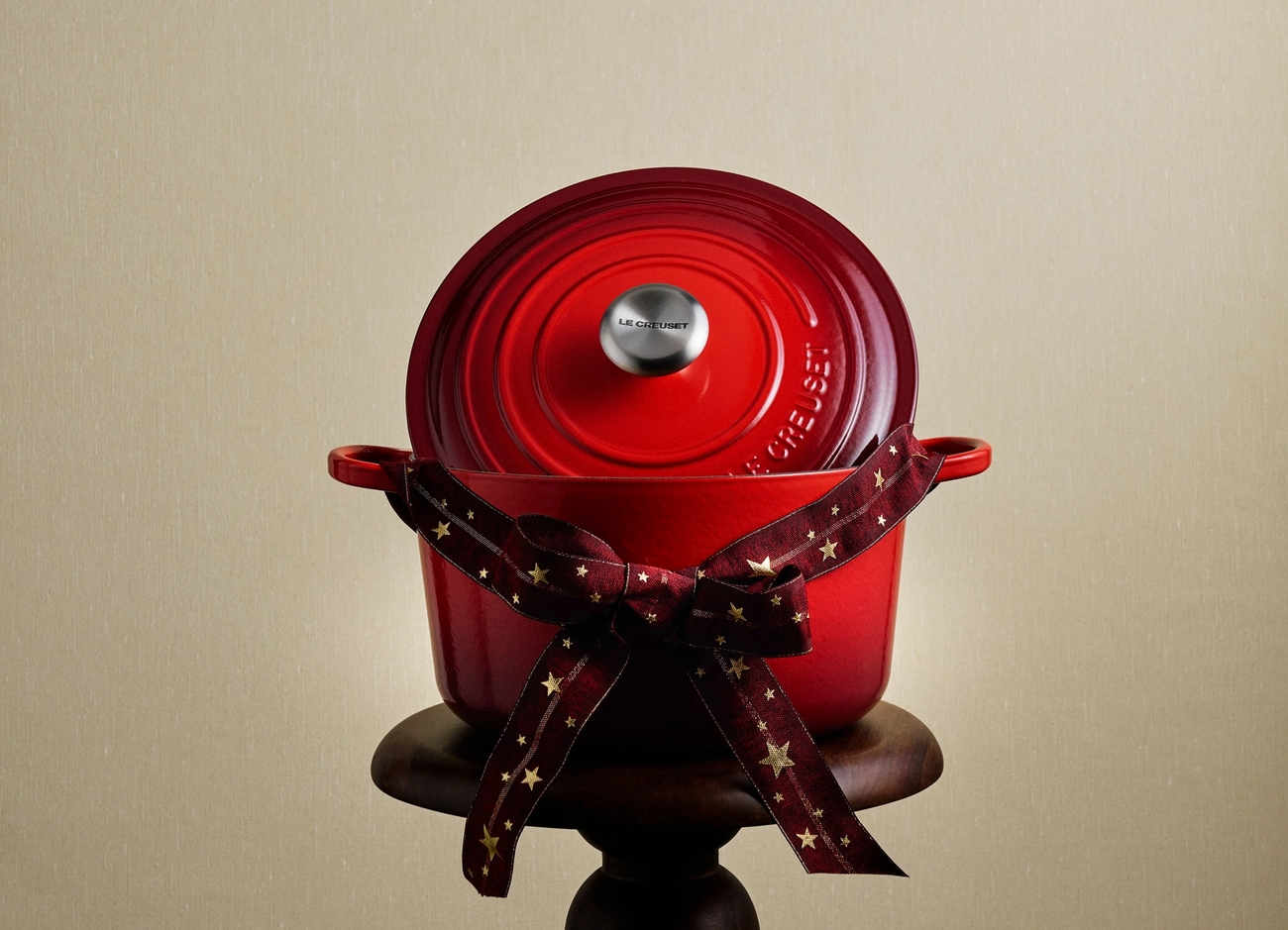 A red Le Creuset Dutch oven, tied with a star-patterned ribbon, sits on a wooden pedestal.