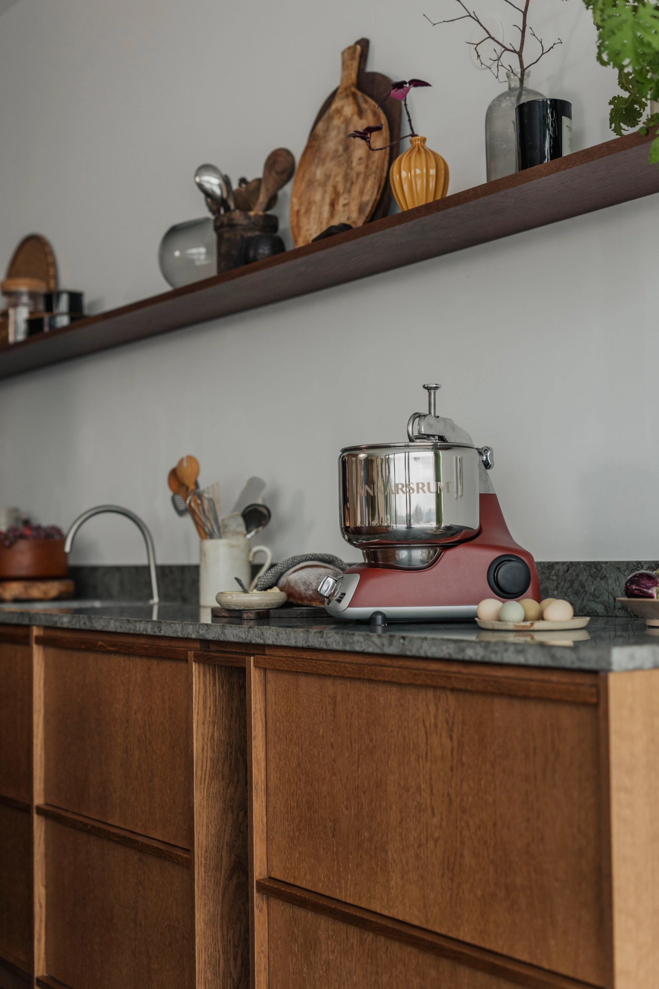 A red stand mixer sits on a speckled granite kitchen counter above wooden cabinets, with a shelf overhead.