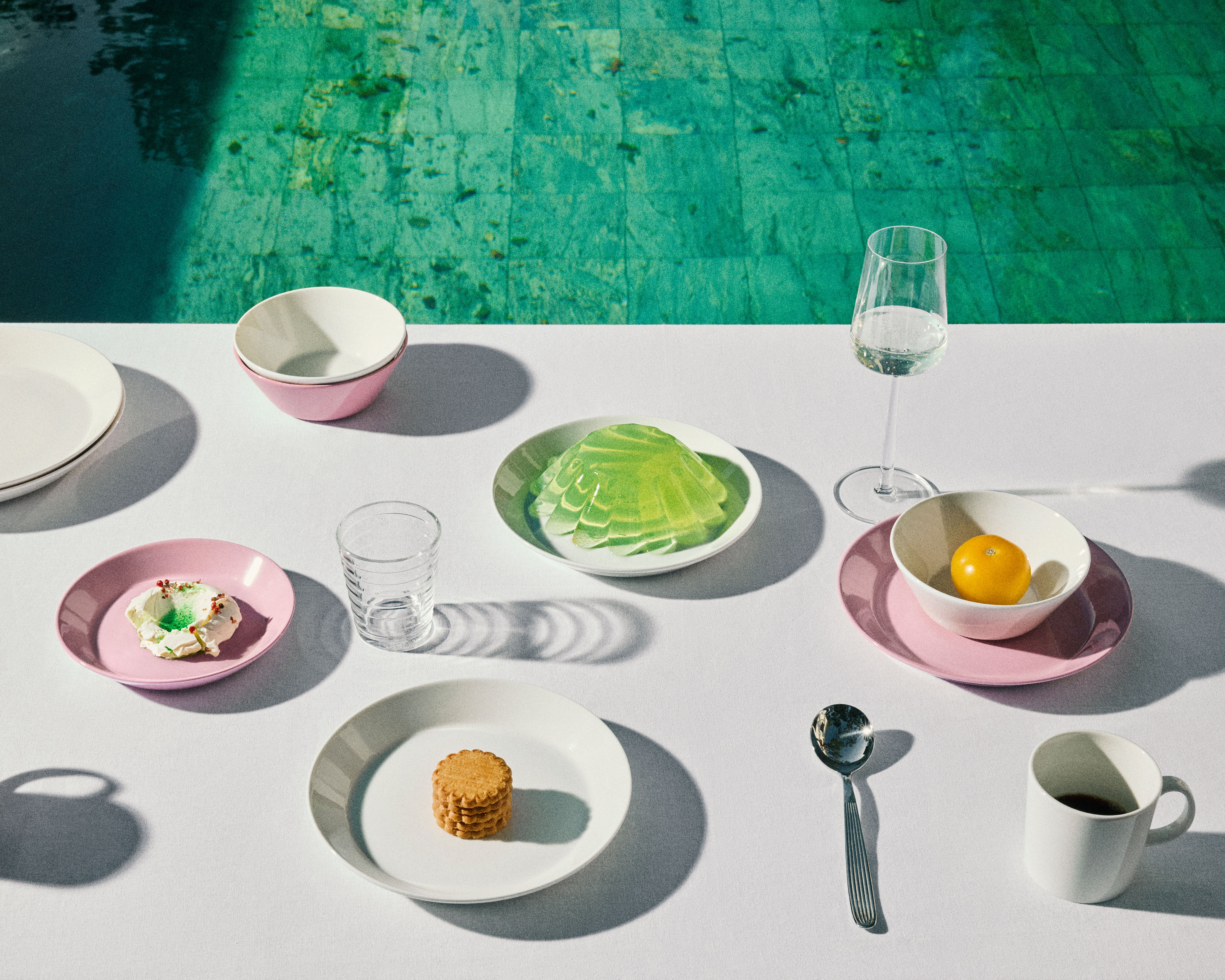 Ett vitt bord med en uppsättning rosa och vita fat med små snacks och drycker, intill en turkos pool.