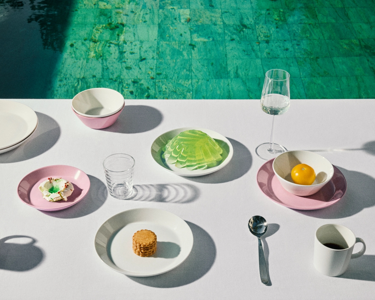 Et hvitt bord med en rekke rosa og hvite tallerkener som inneholder små snacks og drikkevarer, som grenser til et turkis svømmebasseng.