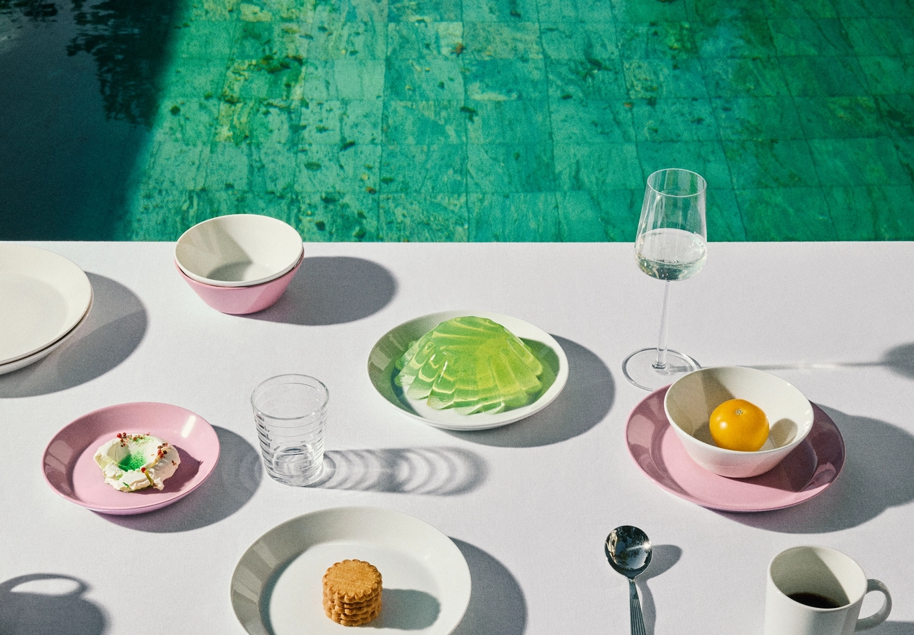 Valkoinen pöytä, jonka päällä on vaaleanpunaisia ja valkoisia astioita, joilla on pieniä välipaloja ja juomia, reunustaen turkoosia uima-allasta.