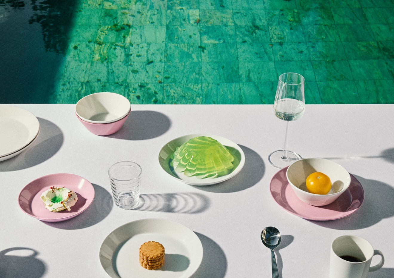 Et hvitt bord med en rekke rosa og hvite tallerkener som inneholder små snacks og drikkevarer, som grenser til et turkis svømmebasseng.