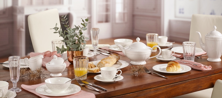 Elegant dukad frukostbord med vitt porslin, croissanter, juice och bröd i ett ljust matsalsrum.