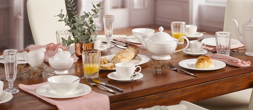 Tyylikkäästi katettu aamiaispöytä, jossa on valkoista posliiniastiastoa, croissanteja, leipää ja mehuja vaaleassa ruokasalissa.