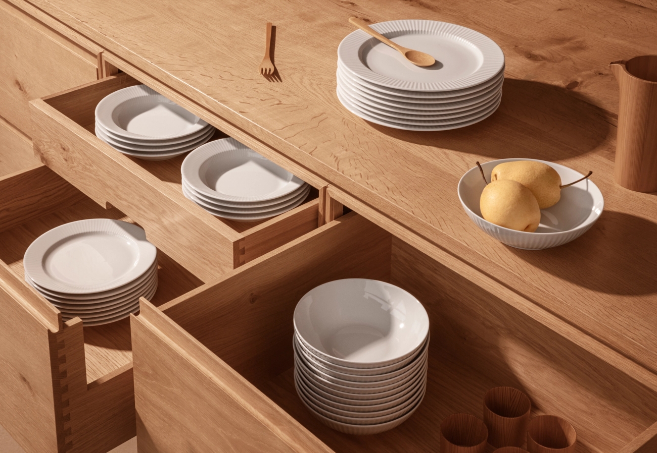 Open wooden kitchen drawers filled with stacks of white plates and bowls, alongside a wooden counter with more plates, pears, and a pitcher.