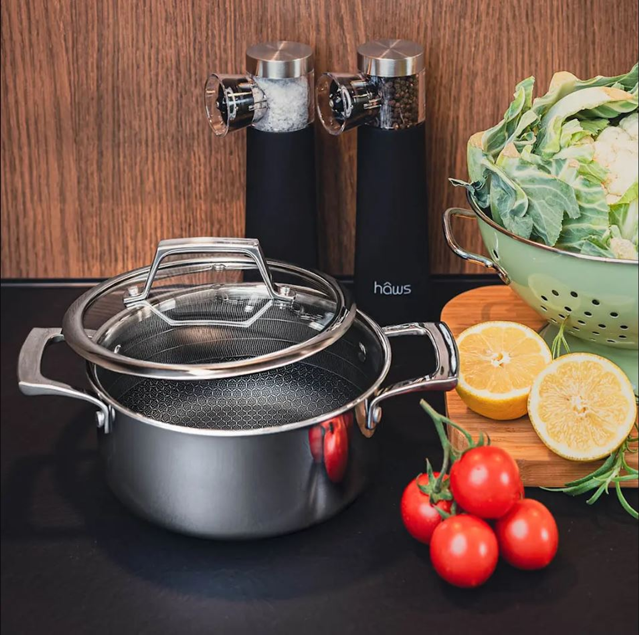 Rostfri kastrull med lock står på en köksbänk bredvid kryddkvarnar, färska tomater, citronhalvor och blomkål i ett durkslag.