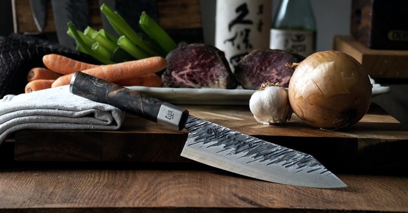 A Japanese chef's knife with a textured blade lies on a wooden board next to raw meat, carrots, green onions, an onion, garlic, and sake.