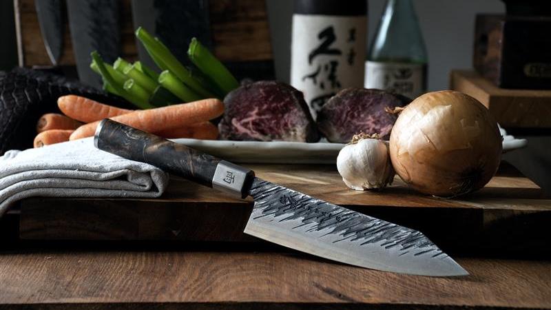 Japanilainen kokkiveitsi ja tuoreet raaka-aineet, kuten sipuli, valkosipuli, porkkanat ja liha, puulla katetulla keittiötasolla.