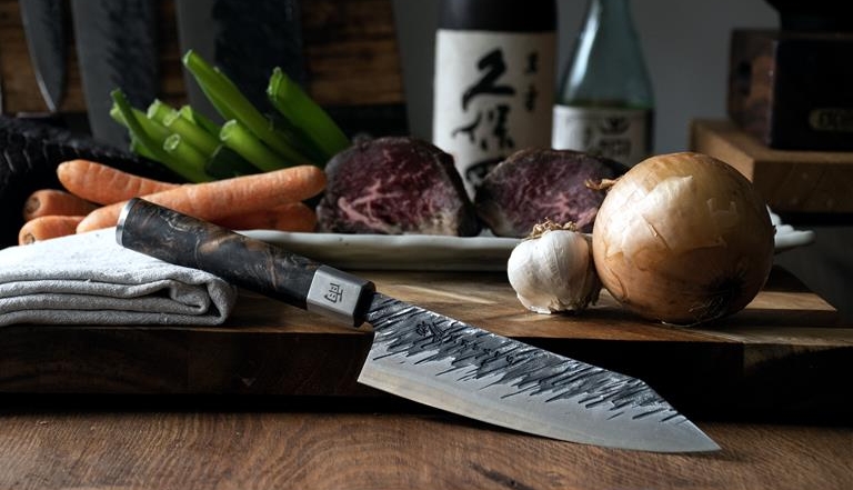 A Japanese kitchen knife with a patterned blade on a wooden board with raw beef, carrots, and vegetables.