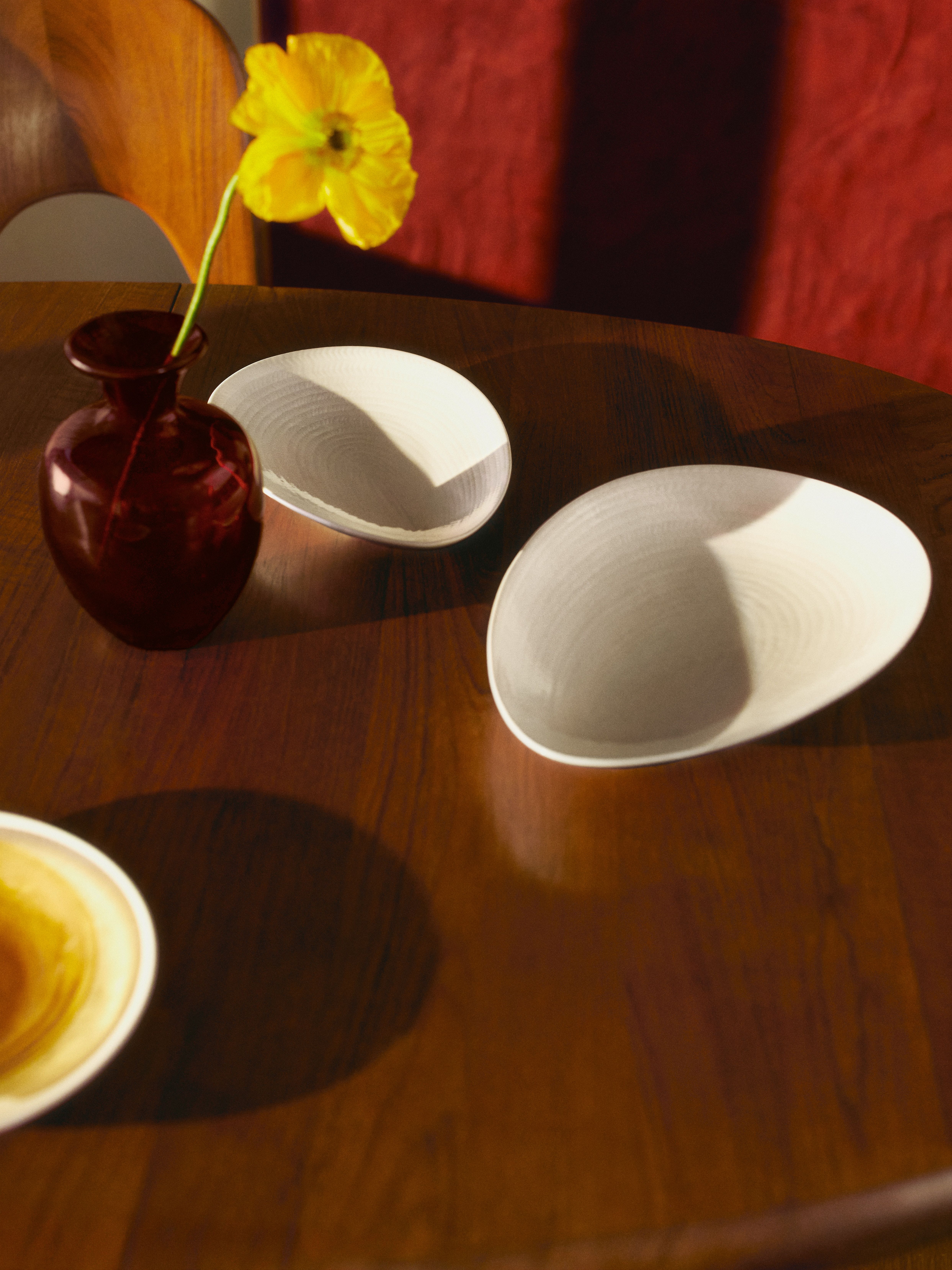 En gul blomma i en röd vas på ett träbord med två ovala, texturerade tallrikar under fläckigt solljus.