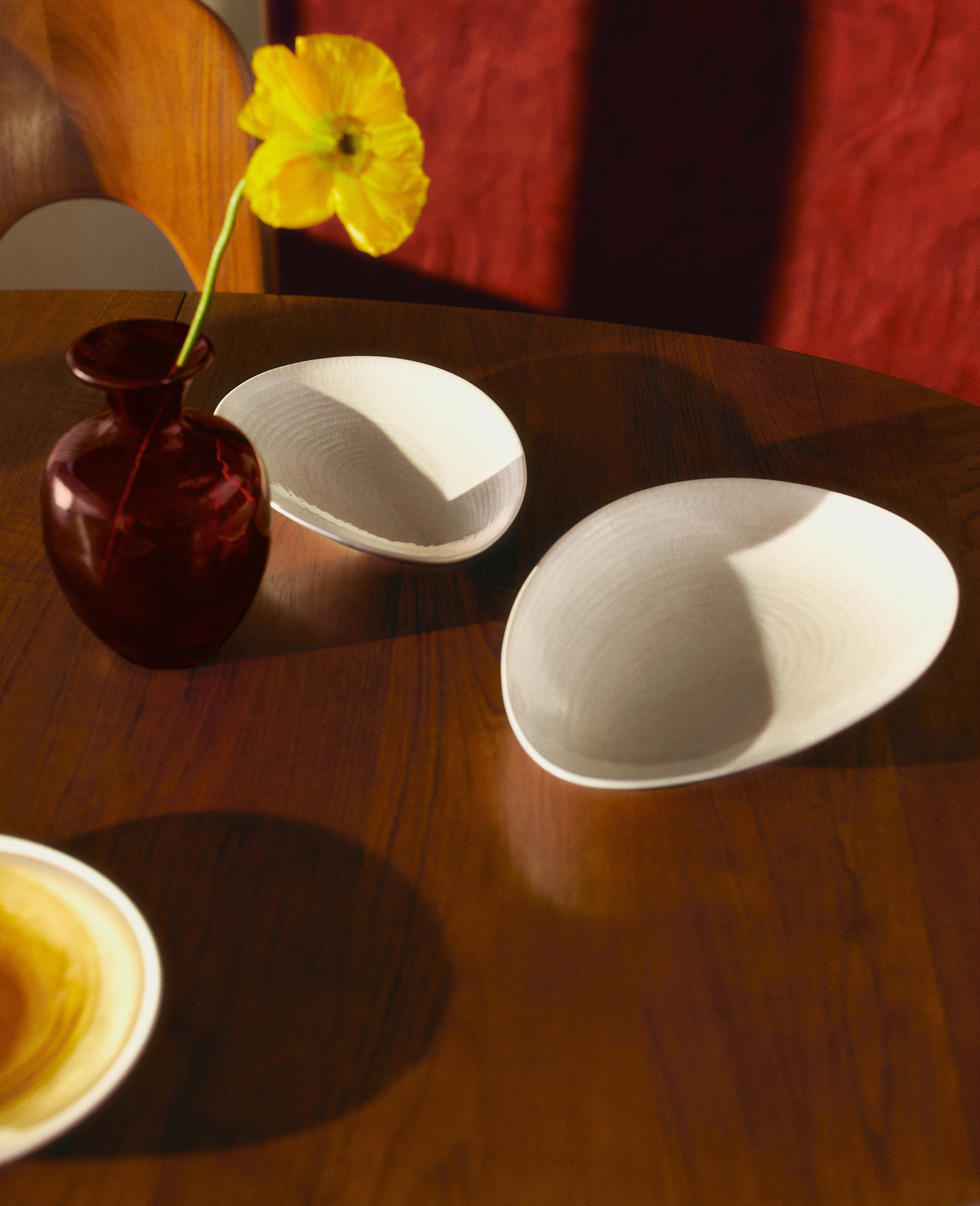 En gul blomma i en röd vas på ett träbord med två ovala, texturerade tallrikar under fläckigt solljus.
