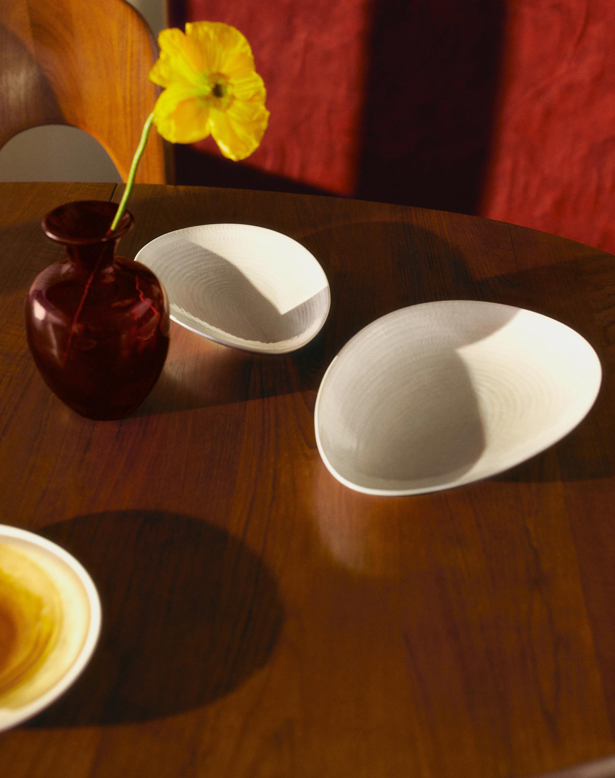 En gul blomma i en röd vas på ett träbord med två ovala, texturerade tallrikar under fläckigt solljus.