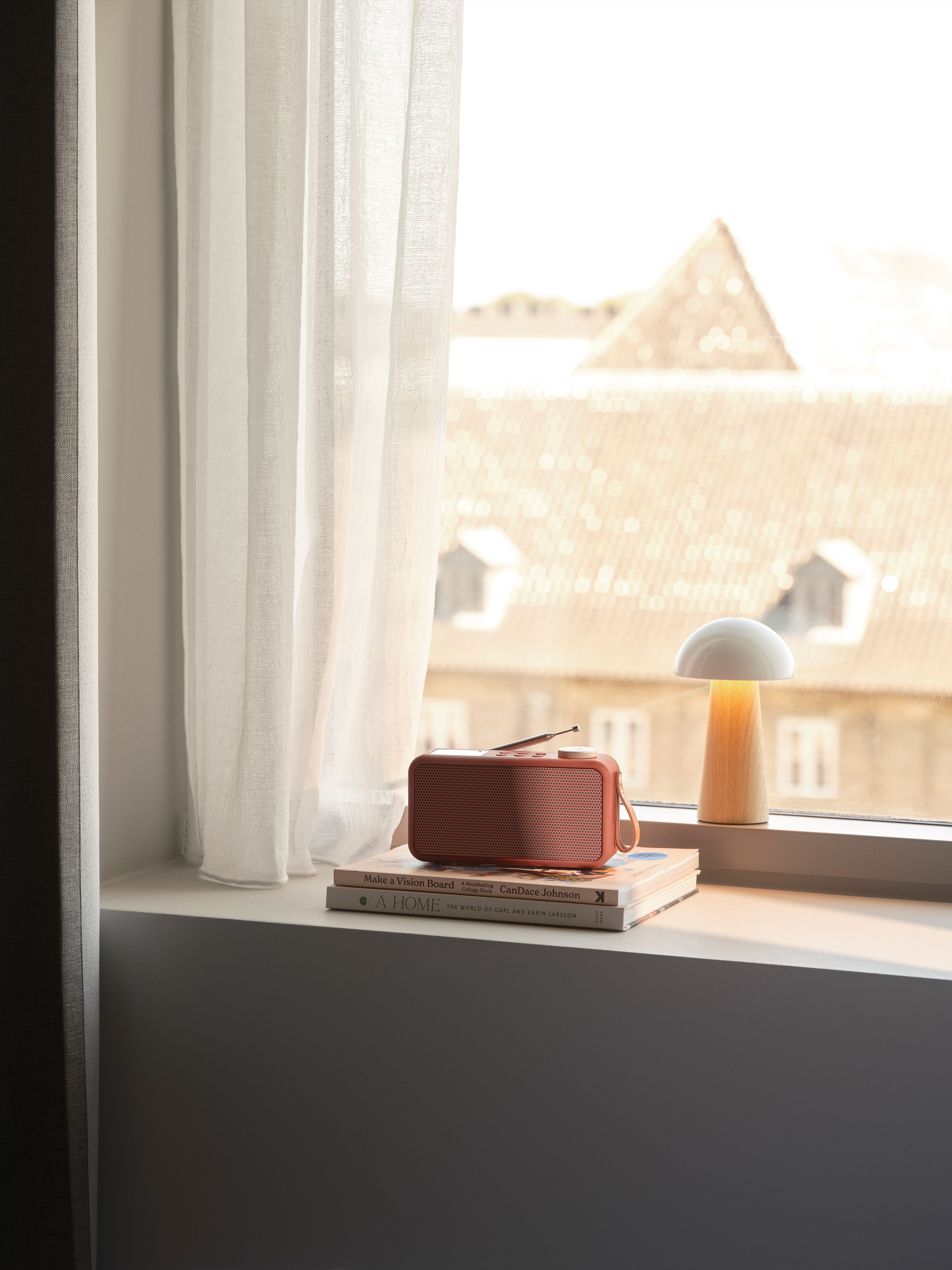 En korallfärgad radio och en lysande svamplampa står på böcker i en solbelyst fönsterkarm, med vita gardiner och en suddig stadsvy i bakgrunden