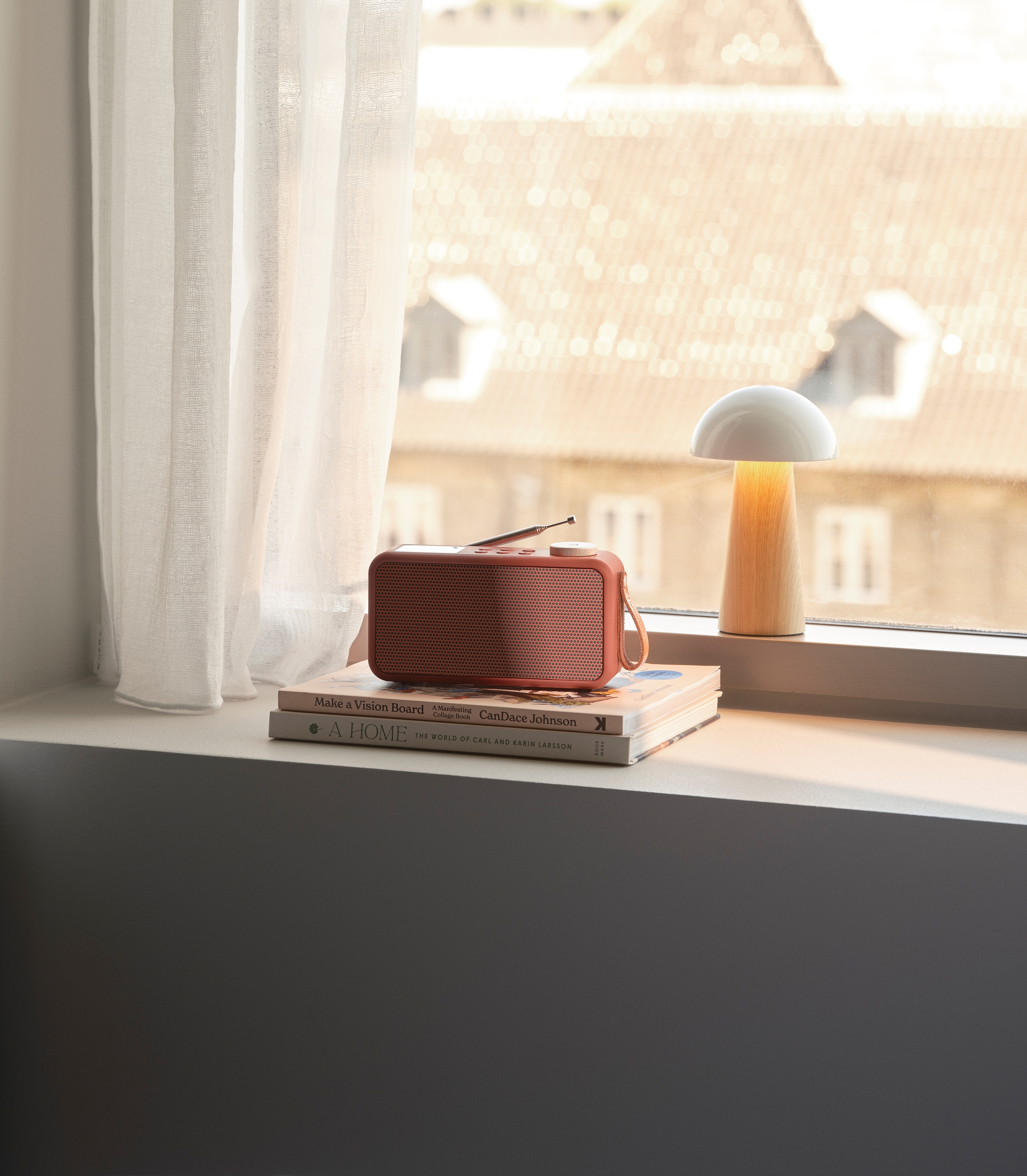 En korallfärgad radio och en lysande svamplampa står på böcker i en solbelyst fönsterkarm, med vita gardiner och en suddig stadsvy i bakgrunden