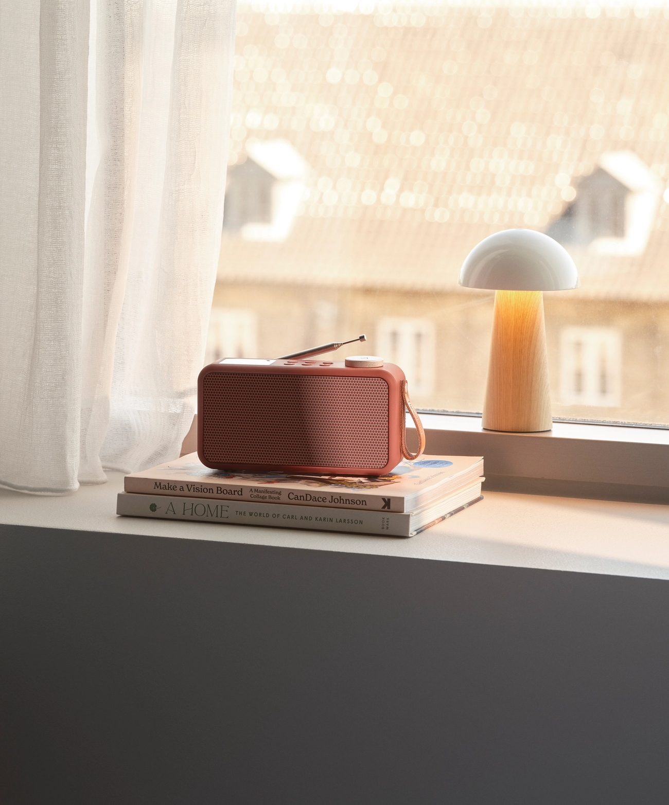 En korallfärgad radio och en lysande svamplampa står på böcker i en solbelyst fönsterkarm, med vita gardiner och en suddig stadsvy i bakgrunden