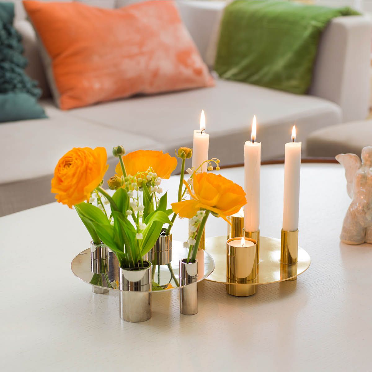 Et sølvfarget blomsterdekor med oransje og hvite blomster ved siden av en gulllysestake med tente hvite lys på et hvitt bord.