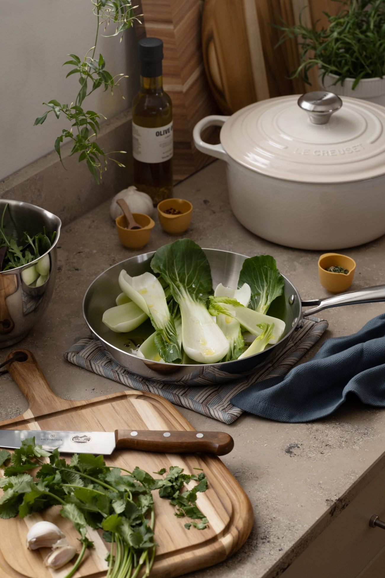 Färsk bok choy i en stekpanna, koriander och vitlök på en skärbräda och köksredskap på en bänkskiva.