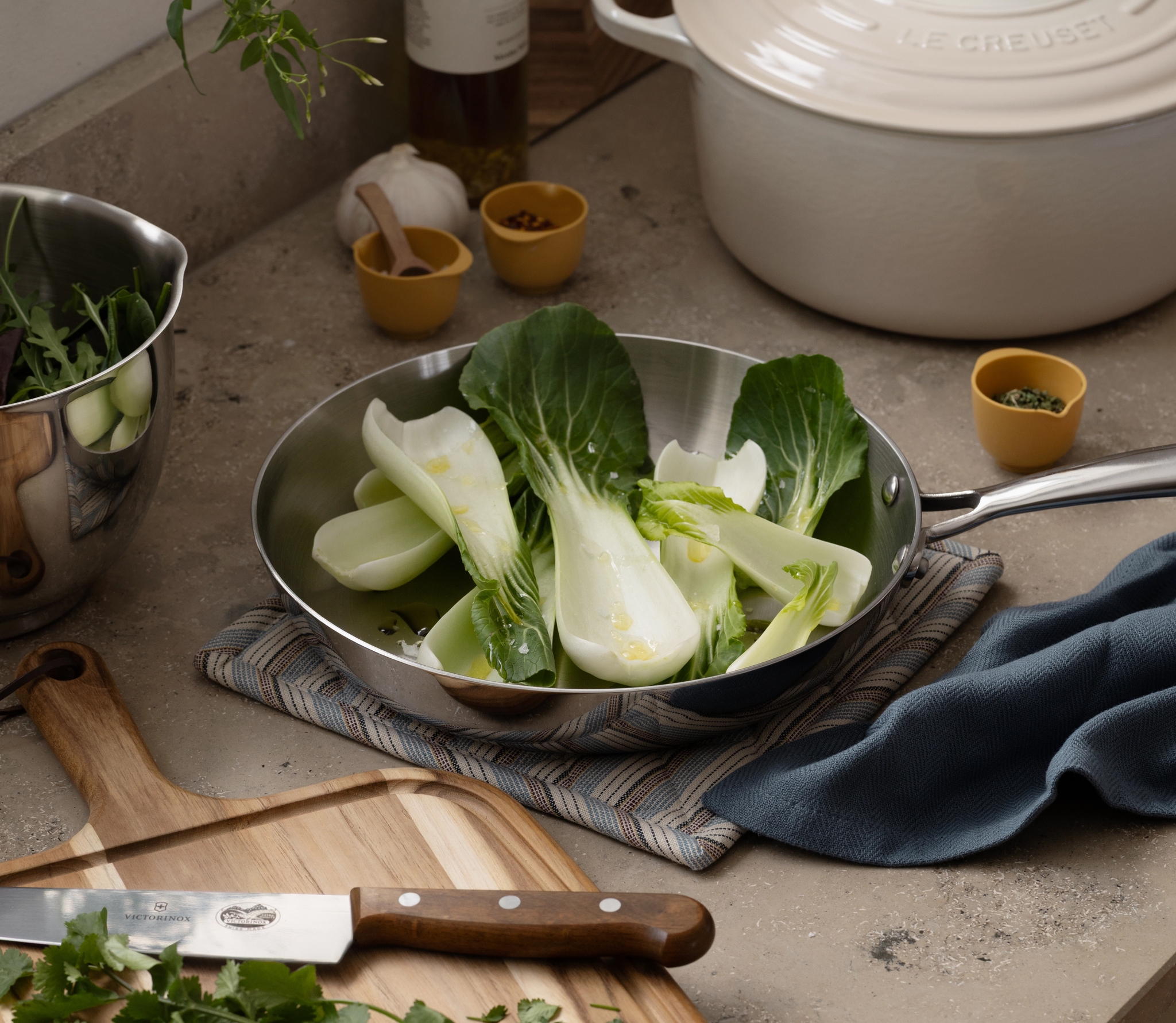 En köksbänk visar bok choy i en stekpanna, en skärbräda med en kniv och örter, en skål med sallad, en vit kastrull och kryddor.