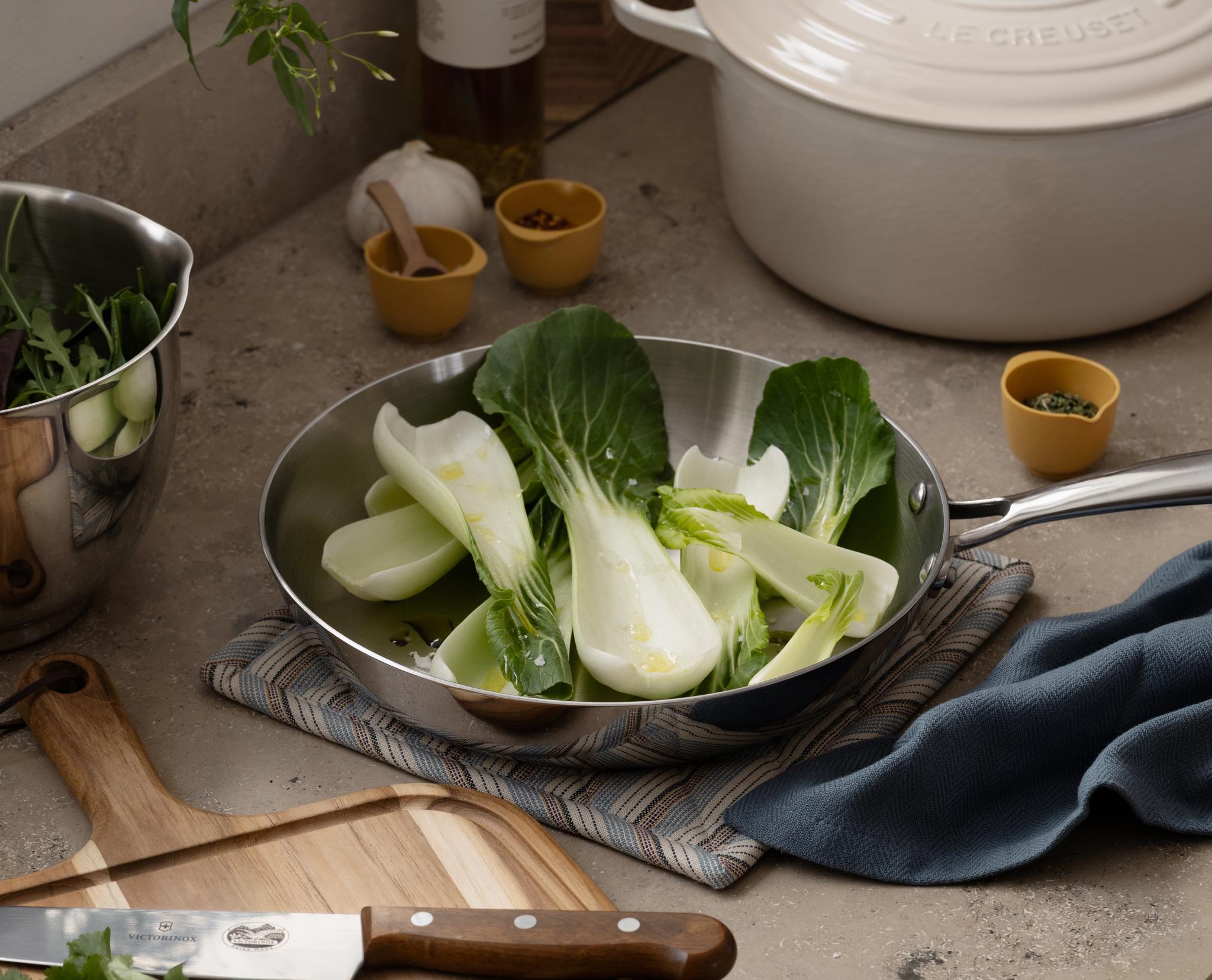 En köksbänk visar bok choy i en stekpanna, en skärbräda med en kniv och örter, en skål med sallad, en vit kastrull och kryddor.