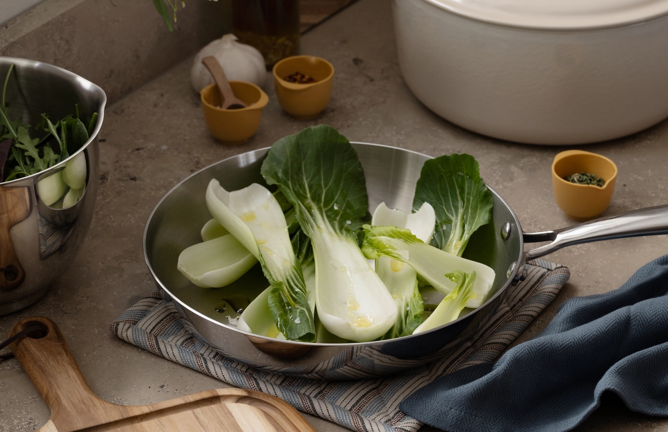 Färsk bok choy i en stekpanna, koriander och vitlök på en skärbräda och köksredskap på en bänkskiva.