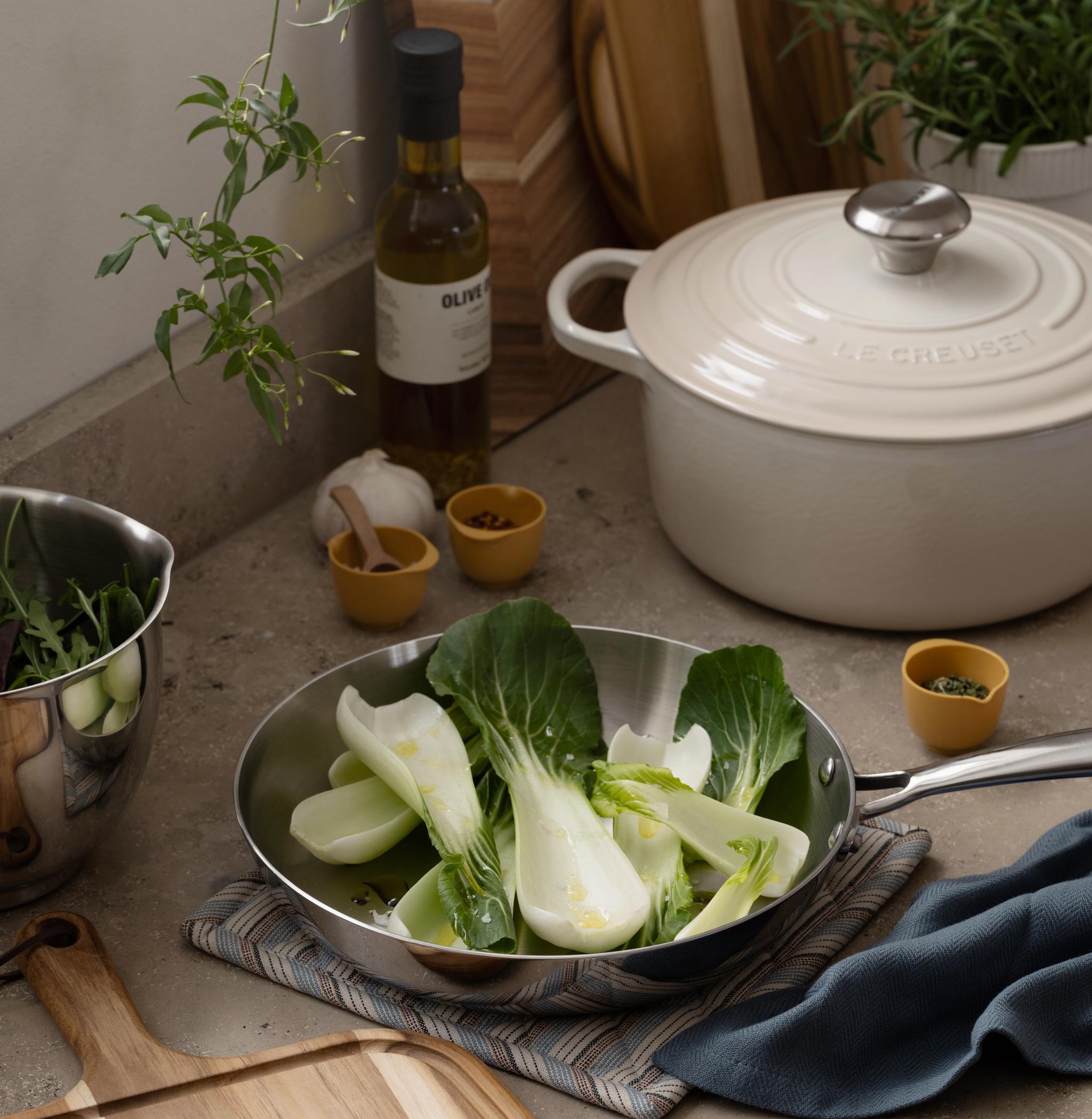 Fresh bok choy in a frying pan on a kitchen counter, with cooking ingredients nearby.