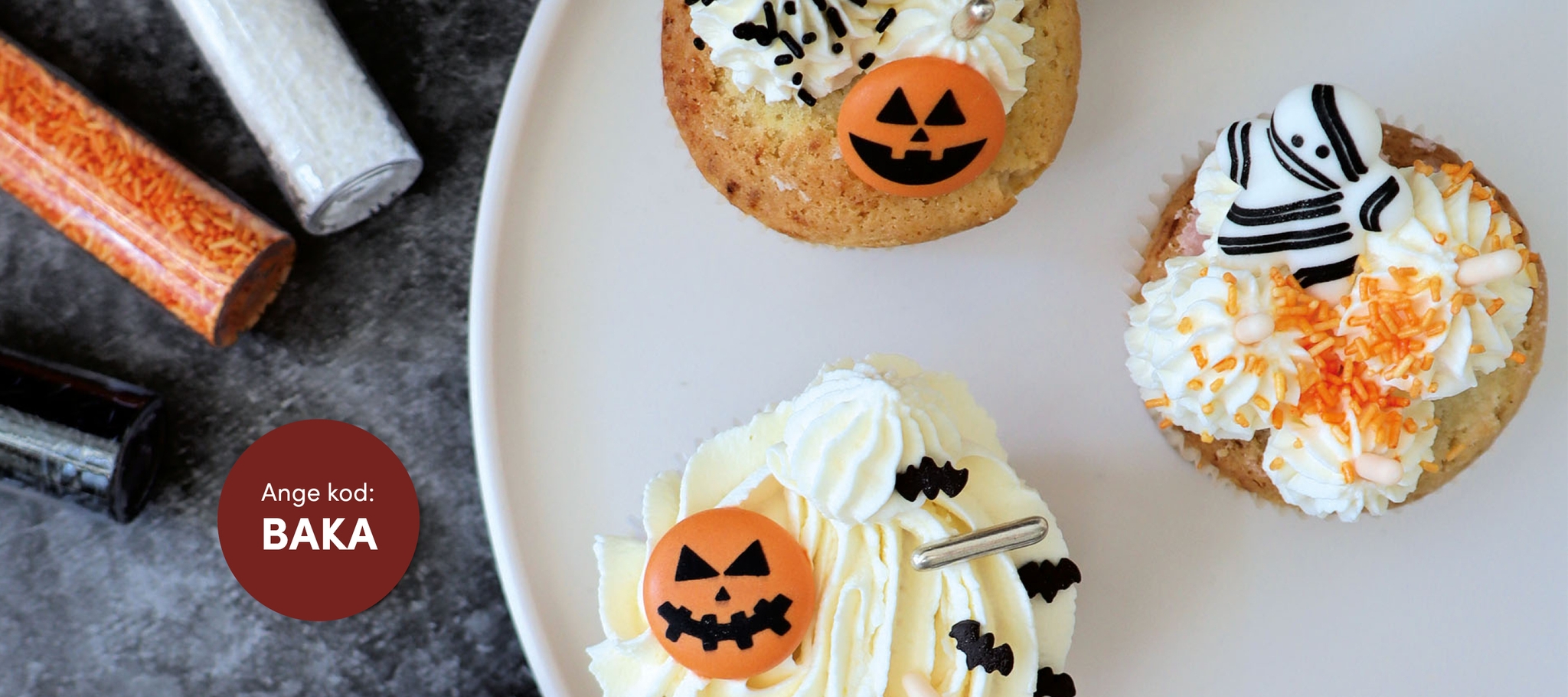 Tre Halloween-cupcakes dekorerade med pumpor, fladdermöss och en mumie ligger på en vit tallrik bredvid tuber med strössel. 
