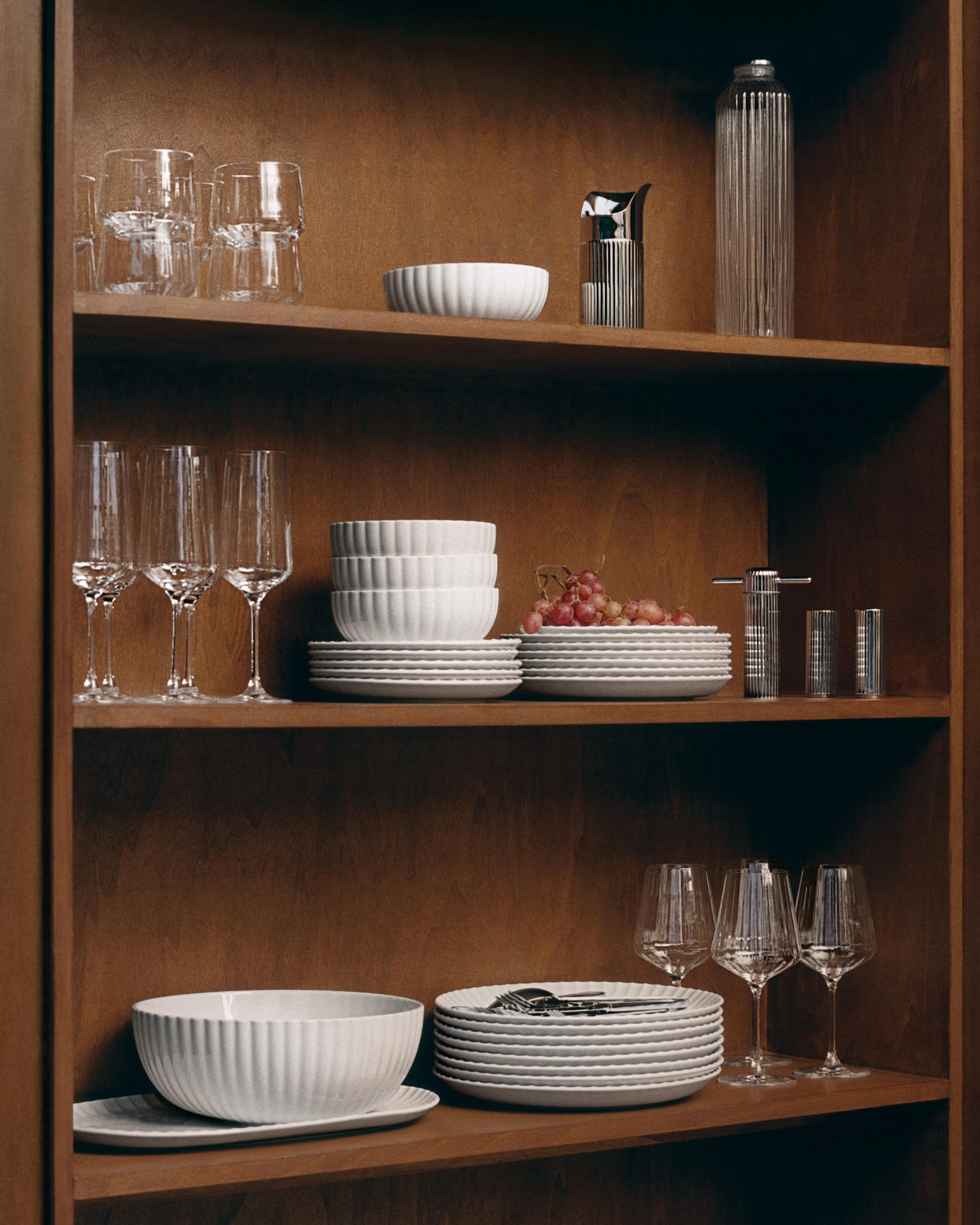 En trähylla med vitt räfflat porslin, klara glas och silverfärgade kökstillbehör.