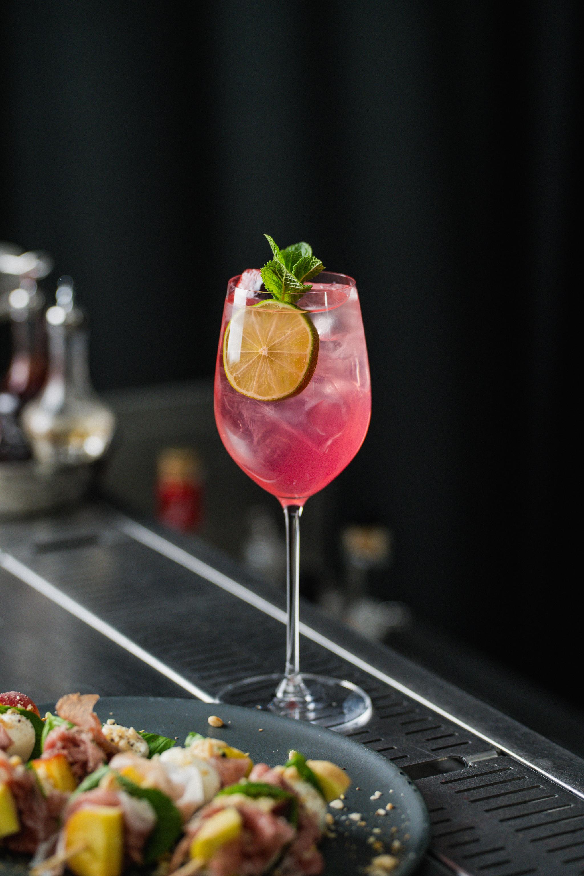 Elegant rosa cocktail med lime- och myntagarnityr serverad vid en bar bredvid en tallrik med plockmat.