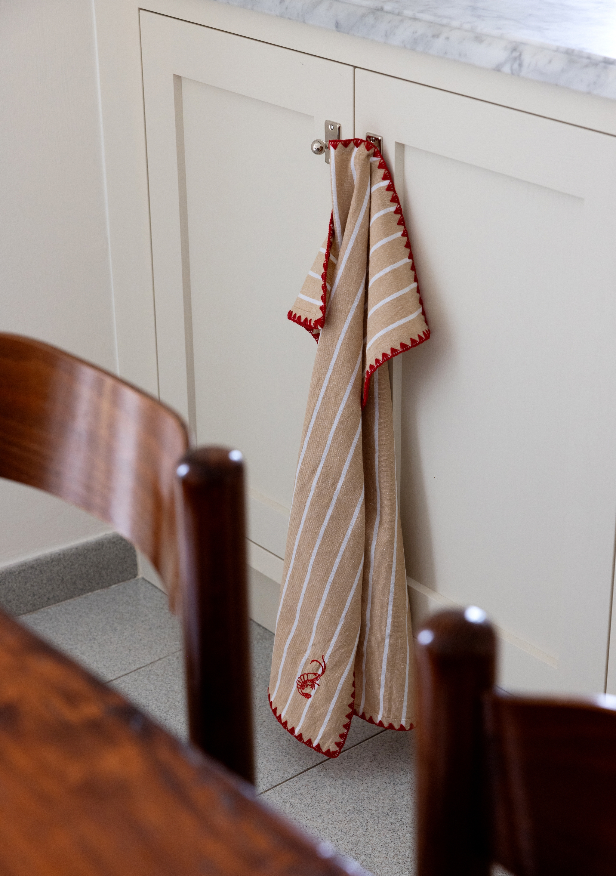 Beige- och vitrandig kökshandduk med röd kant hänger på ett vitt köksskåp.