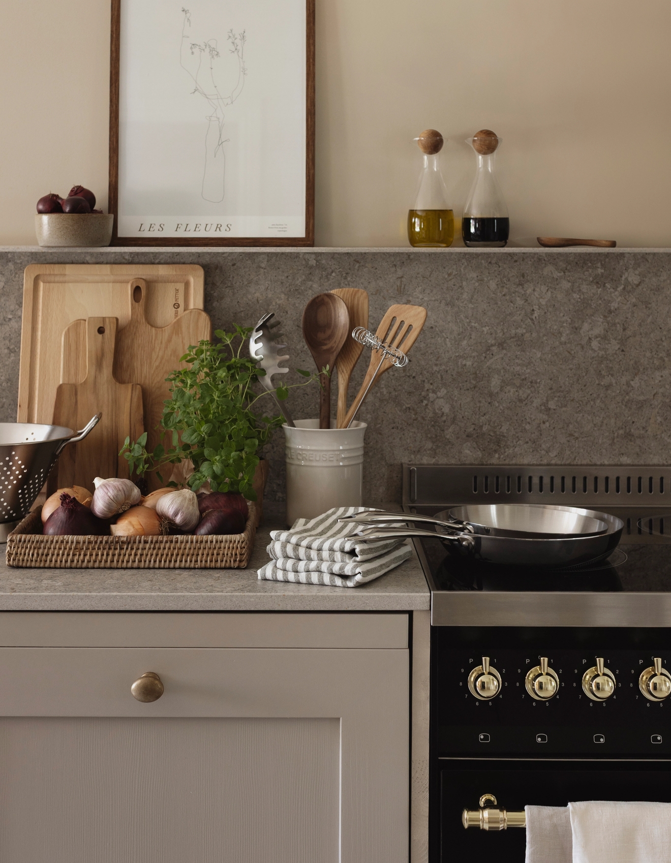 A rustic kitchen counter with a framed floral print, cooking oil, various cutting boards, fresh herbs, vegetables, utensils, and a frying pan on a black stovetop.