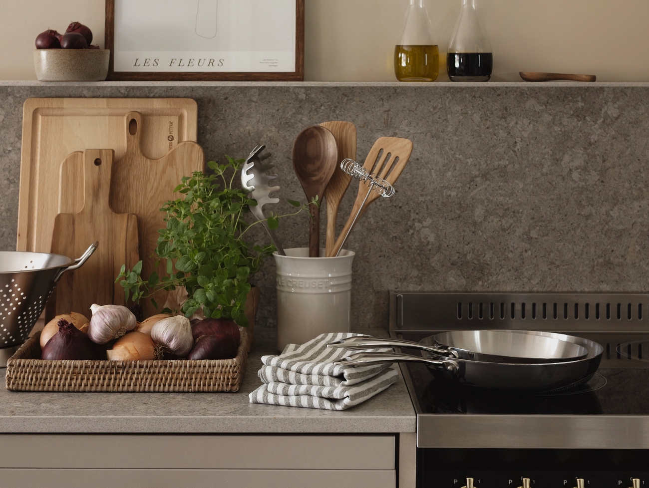 Stilren köksbänk i beige med kryddväxt, lök, skärbrädor, stekpanna och oljeflaskor mot en varm, neutral vägg.
