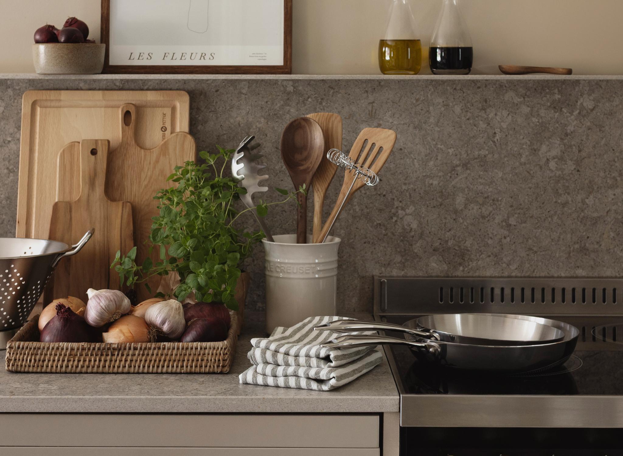 Stilren kjøkkenbenk med urter, løk, skjærefjøler og stekepanne ved siden av en komfyr i moderne landlig stil.