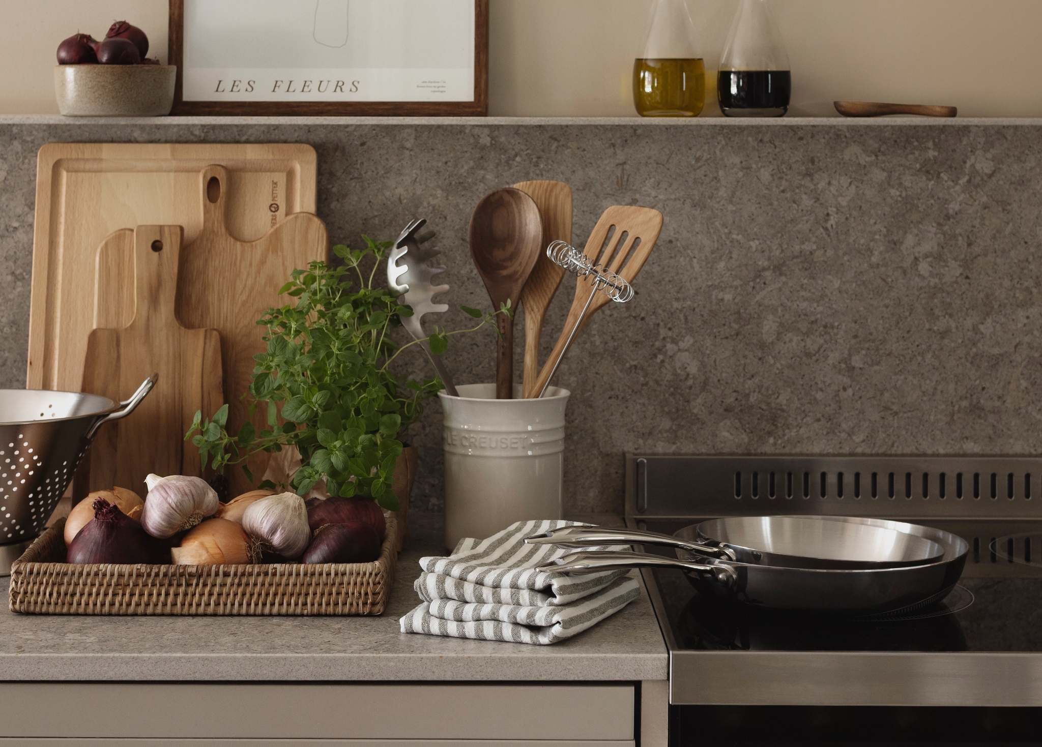 Stilren kjøkkenbenk med urter, løk, skjærefjøler og stekepanne ved siden av en komfyr i moderne landlig stil.