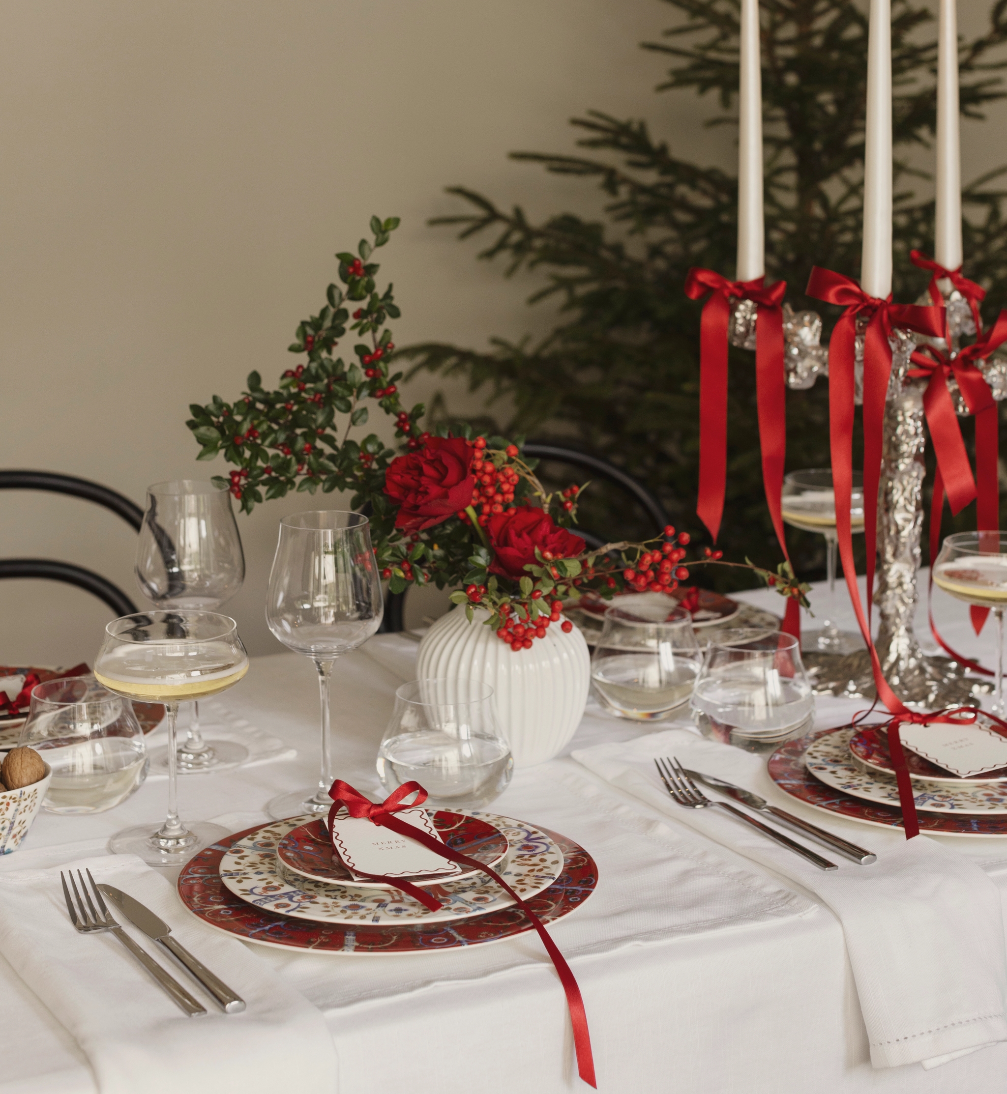 Juhlava jouluinen ruokapöytä, jossa on punakuvioiset lautaset, punaisista ruusuista ja marjoista koostuva pöytäasetelma sekä hopeinen kynttelikkö, jossa on sytytettyjä valkoisia kynttilöitä ja punaisia nauhoja, taustalla joulukuusi.