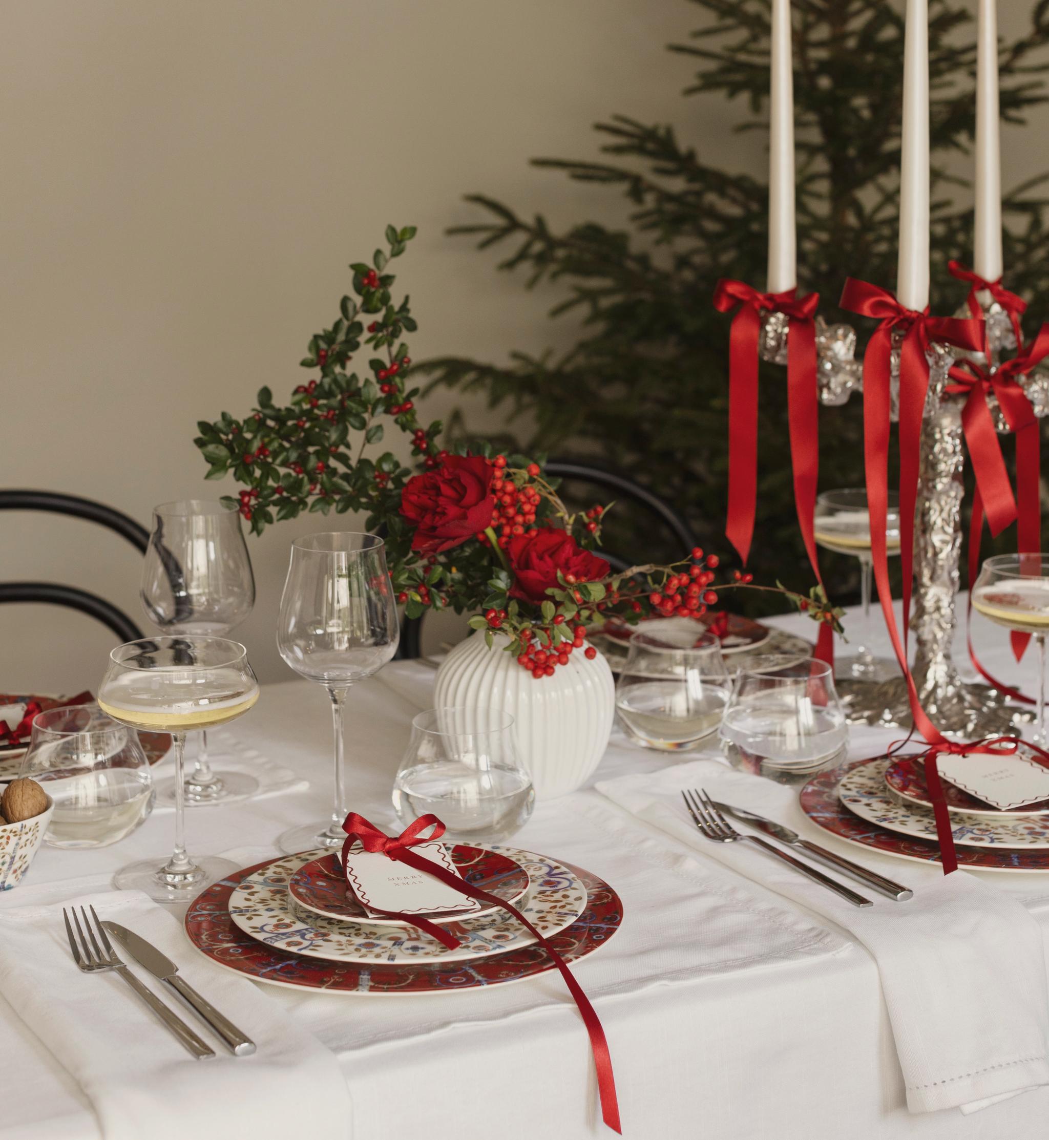 Juhlava jouluinen ruokapöytä, jossa on punakuvioiset lautaset, punaisista ruusuista ja marjoista koostuva pöytäasetelma sekä hopeinen kynttelikkö, jossa on sytytettyjä valkoisia kynttilöitä ja punaisia nauhoja, taustalla joulukuusi.