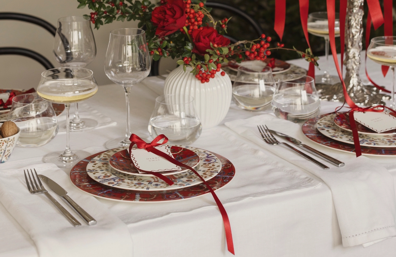 Elegant julbord med vitt linne, röda och mönstrade tallrikar, bestick, champagneglas och en röd blommig mittdekoration.