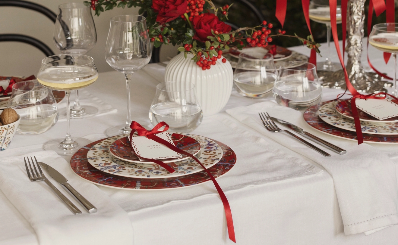 Elegant julbord med vitt linne, röda och mönstrade tallrikar, bestick, champagneglas och en röd blommig mittdekoration.