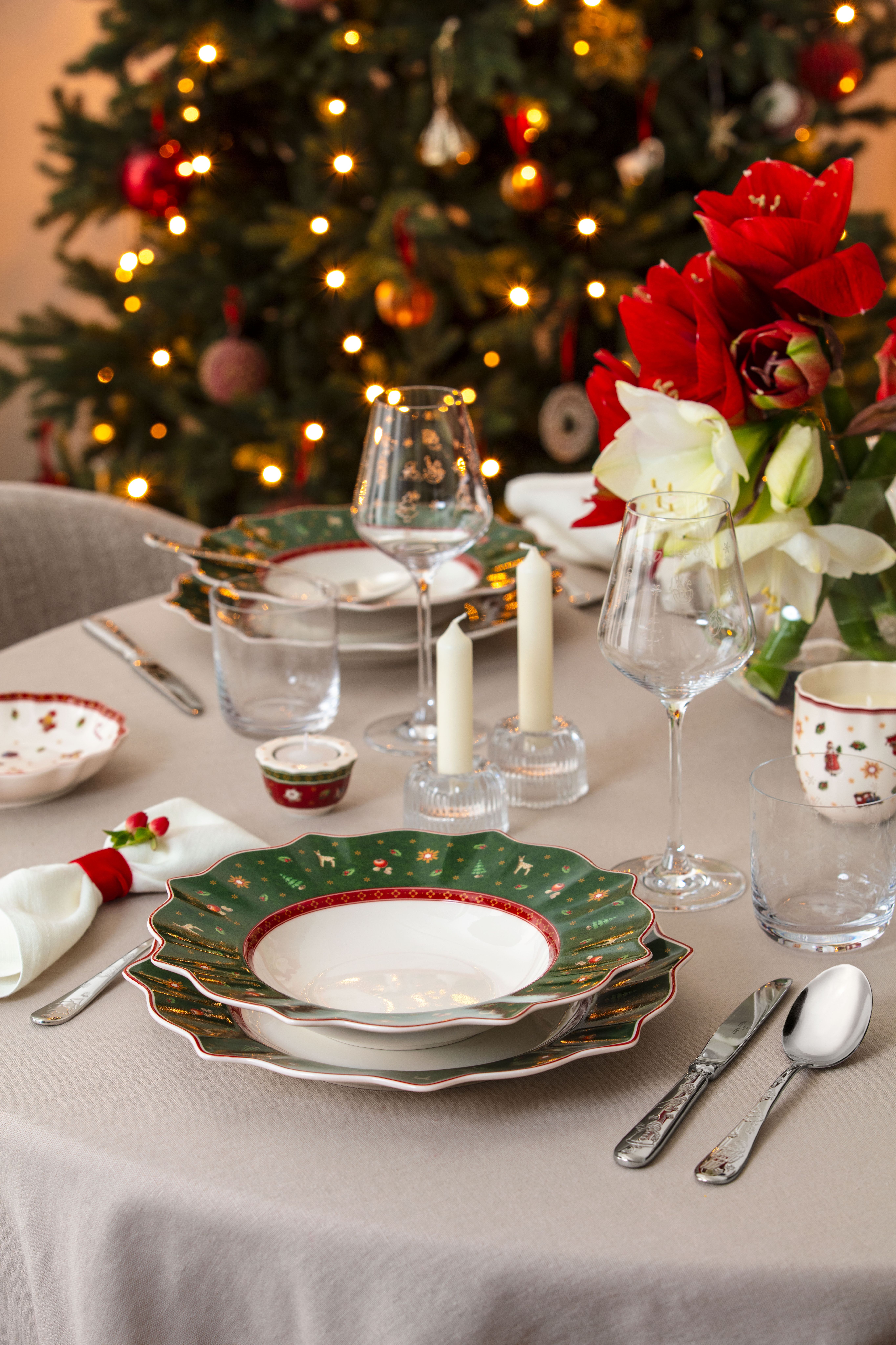 Et julebord dekket med grønne mønstrede tallerkener, røde og hvite blomster, lys og et blinkende juletre i bakgrunnen.