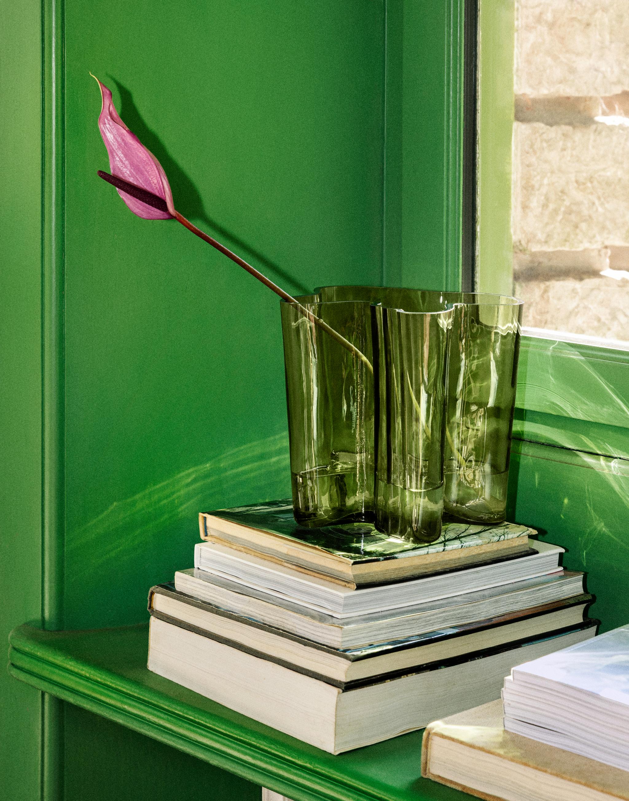 En livfullt rosa anthuriumblomma i en grön glasvas står på en stapel böcker på en klargrön hylla