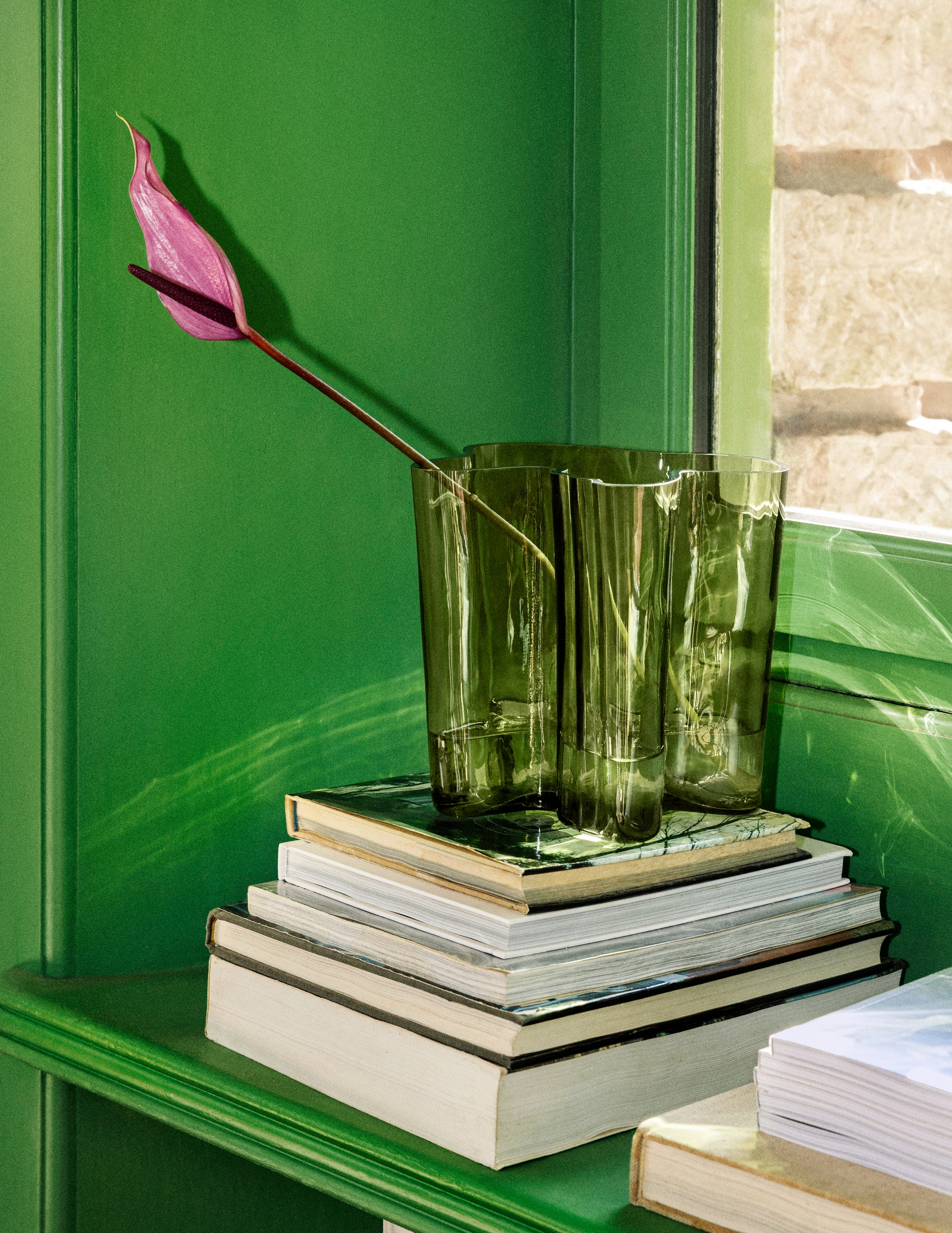 En livfullt rosa anthuriumblomma i en grön glasvas står på en stapel böcker på en klargrön hylla