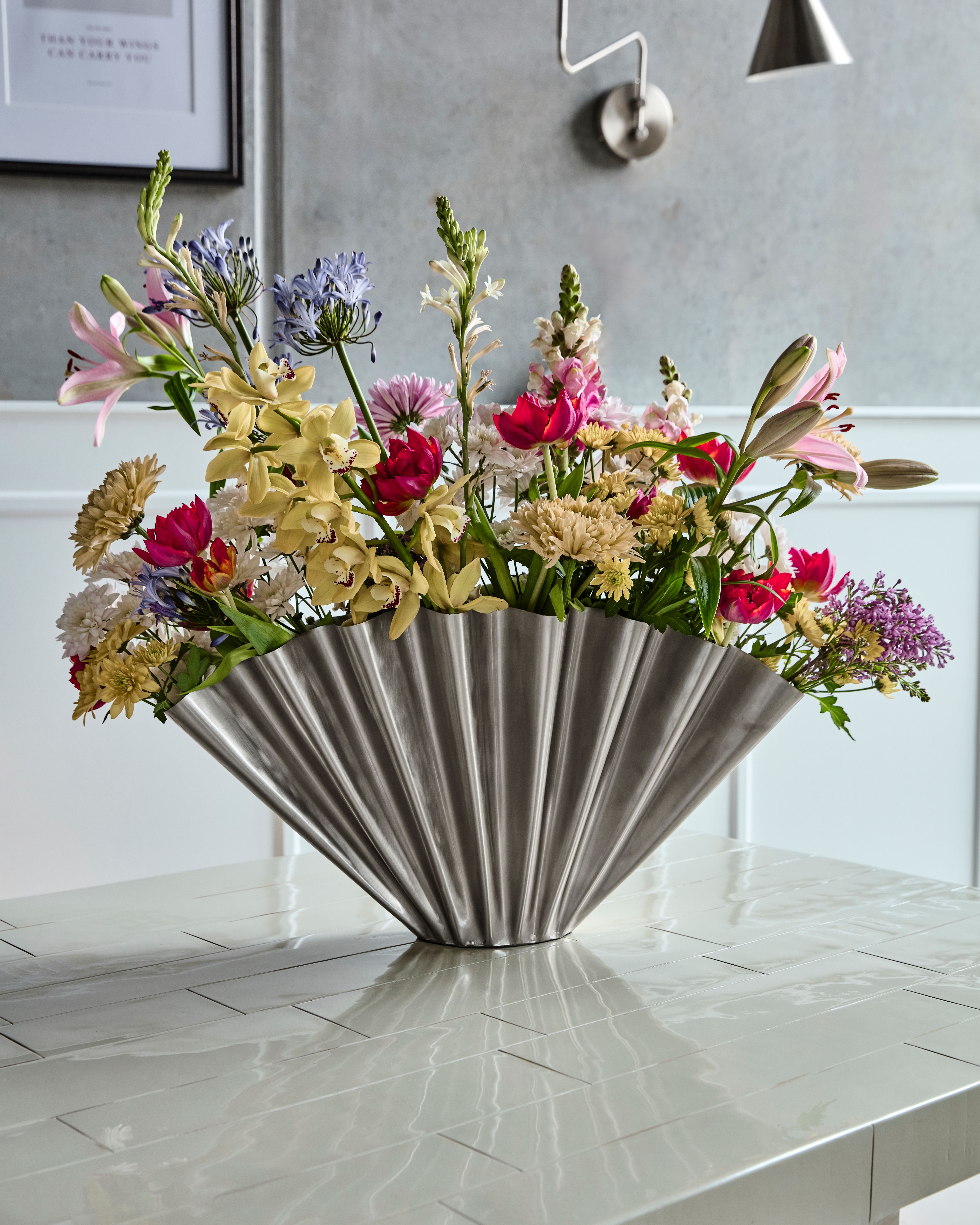 A large, colorful floral arrangement in a silver fan-shaped vase sits on a glossy tiled table.