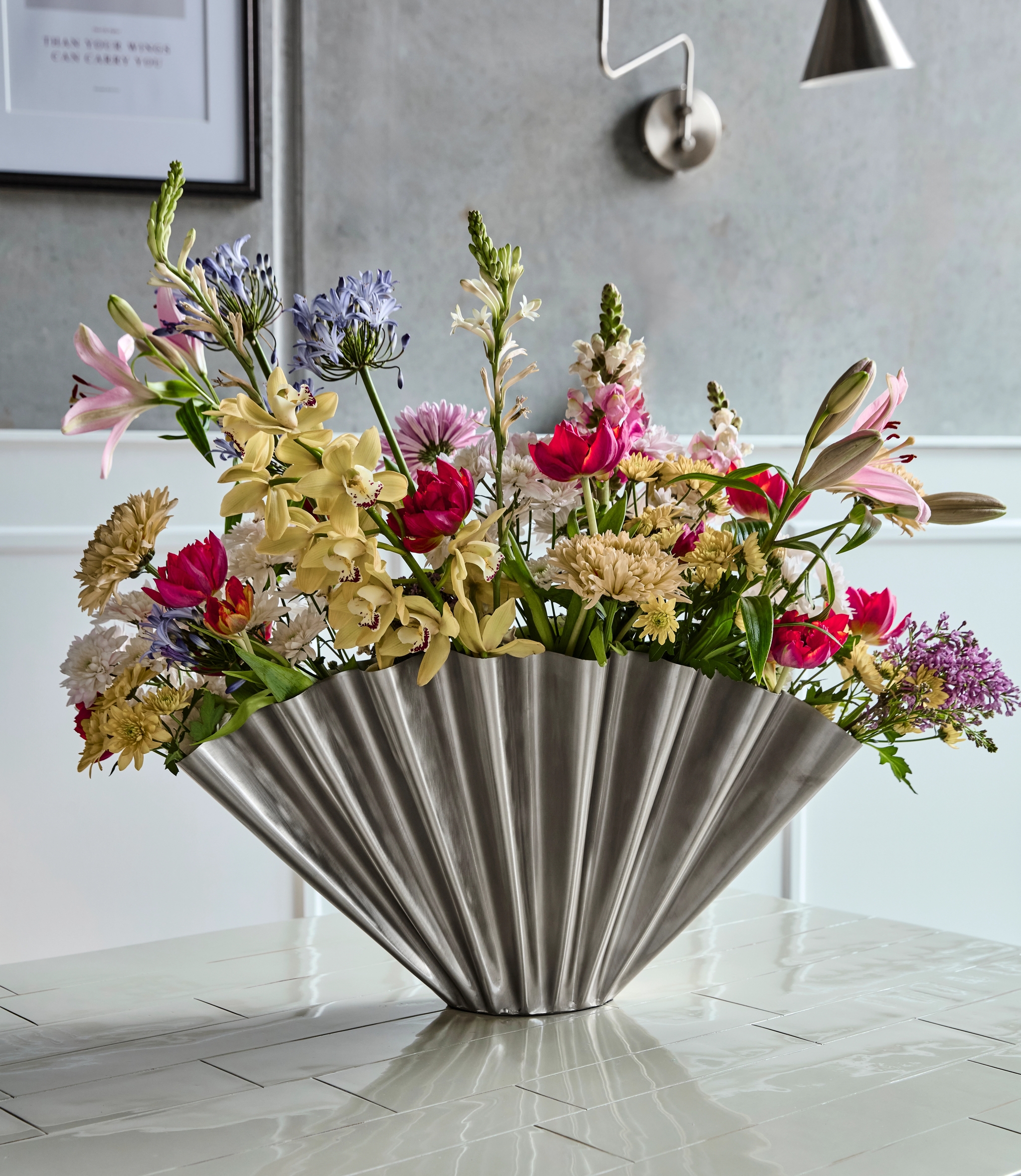 A large, colorful floral arrangement in a silver, ribbed fan-shaped vase on a white tiled table.