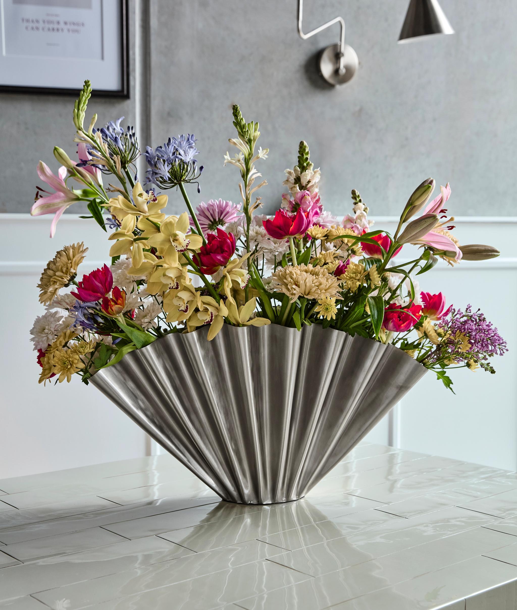A large, colorful floral arrangement in a silver fan-shaped vase sits on a glossy tiled table.