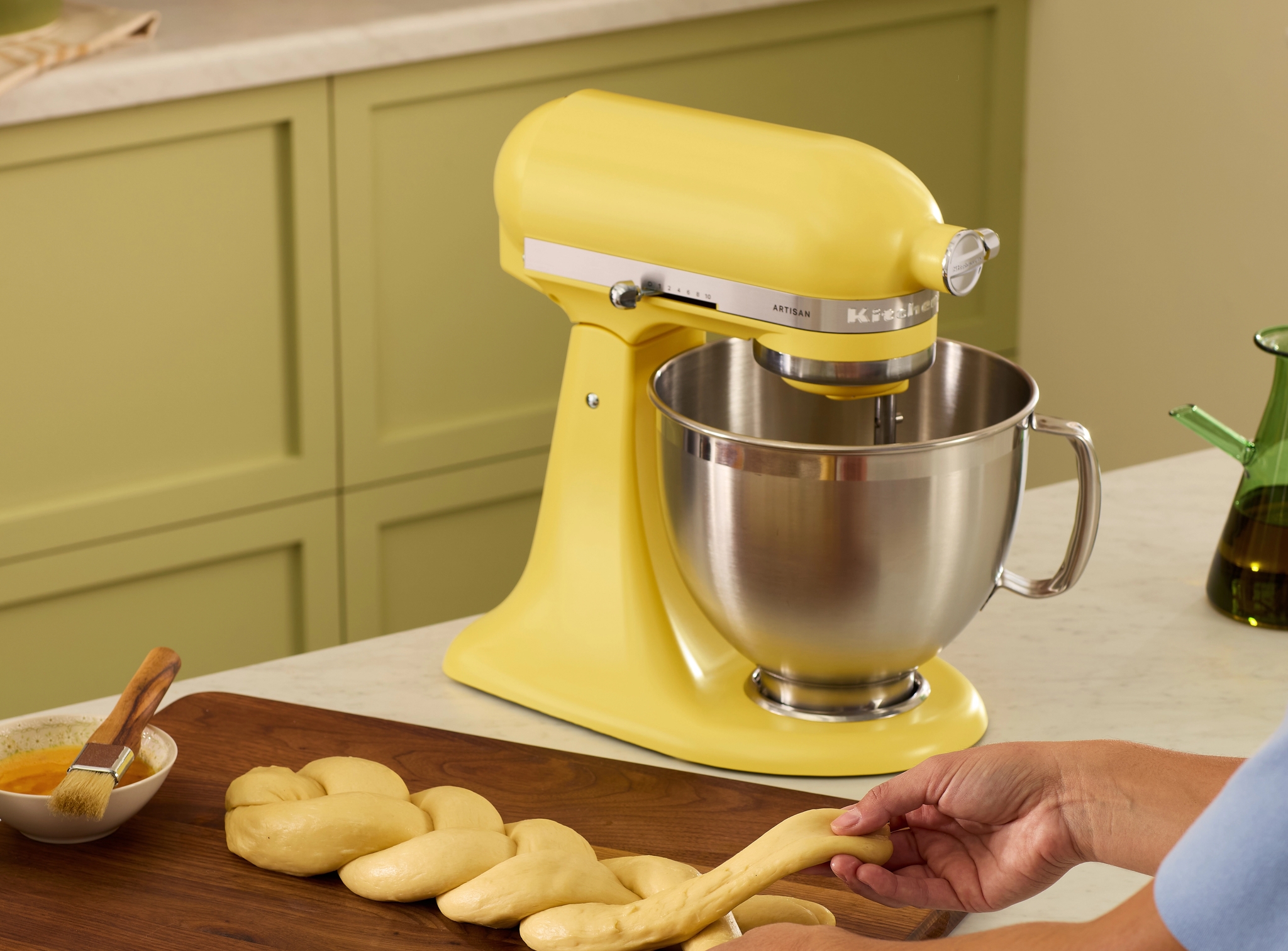 Henkilö letittää taikinaa puisella leikkuulaudalla keltaisen KitchenAid-yleiskoneen vieressä.