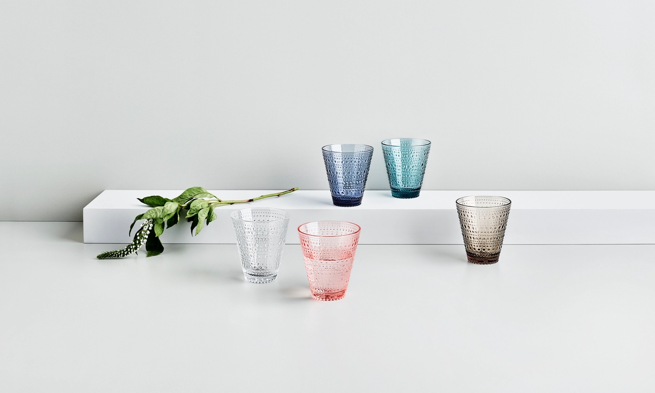 Drikkeglass Iittala Kastehelmi i ulike farger, samt grønn kvist mot hvit bakgrunn