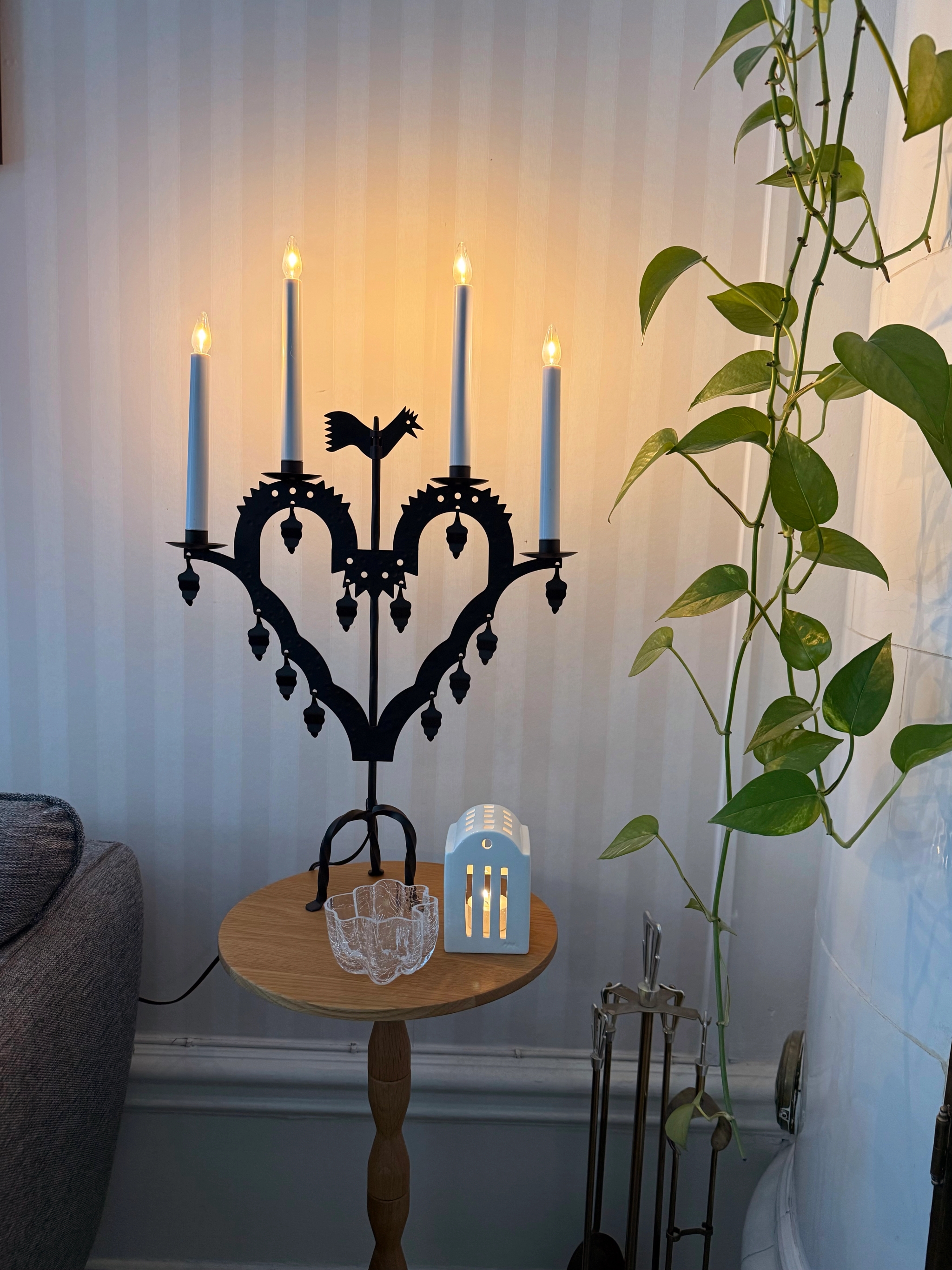 A black heart-shaped candelabra with a rooster and four glowing electronic candles sits on a round wooden table next to a white house-shaped lantern and a glass dish, with a tall trailing green plant on the right.