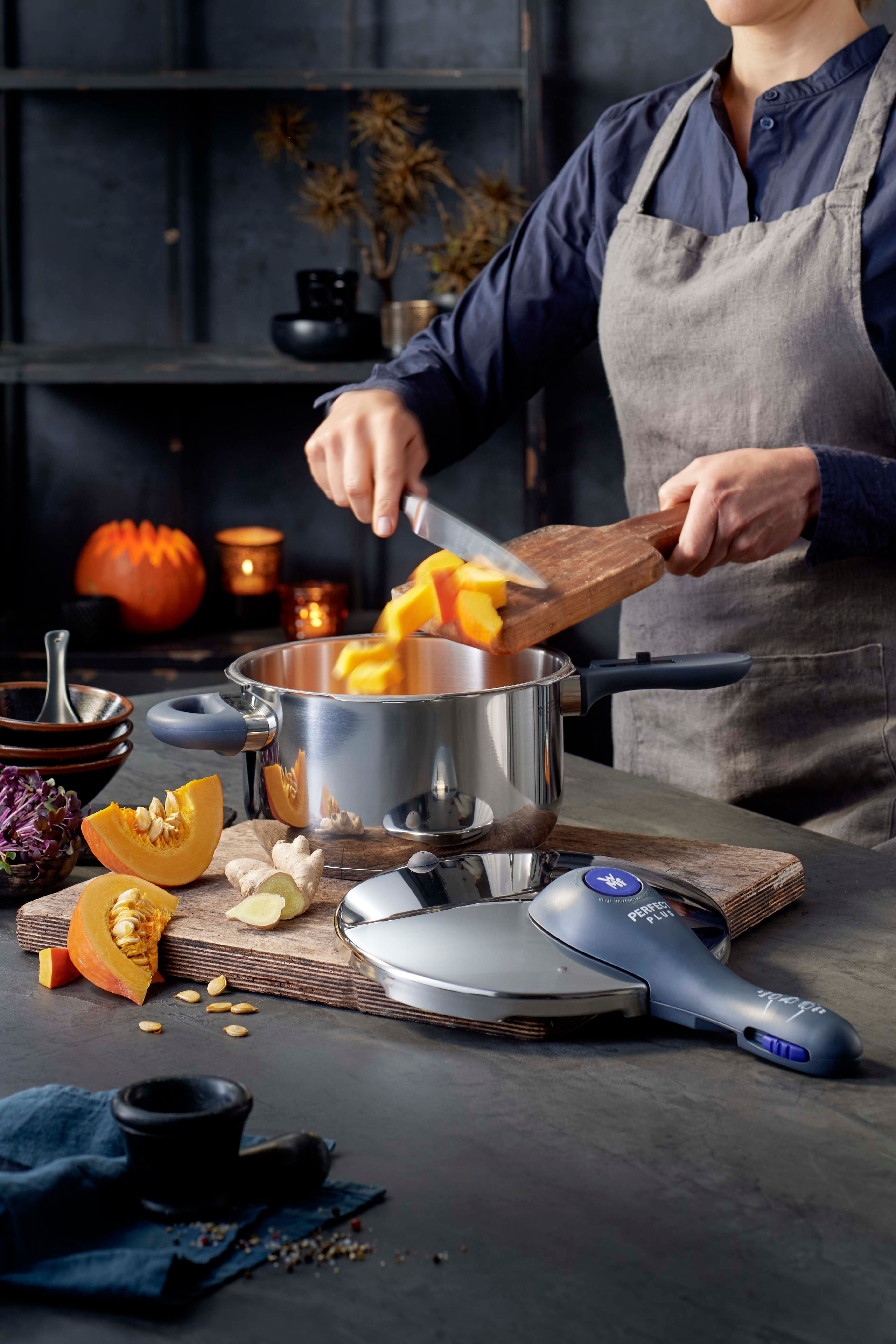 Lähikuva kokin käsivarresta puisella keittiön tasolla vasaroitujen ruostumattomasta teräksestä valmistettujen keittoastioiden vieressä.