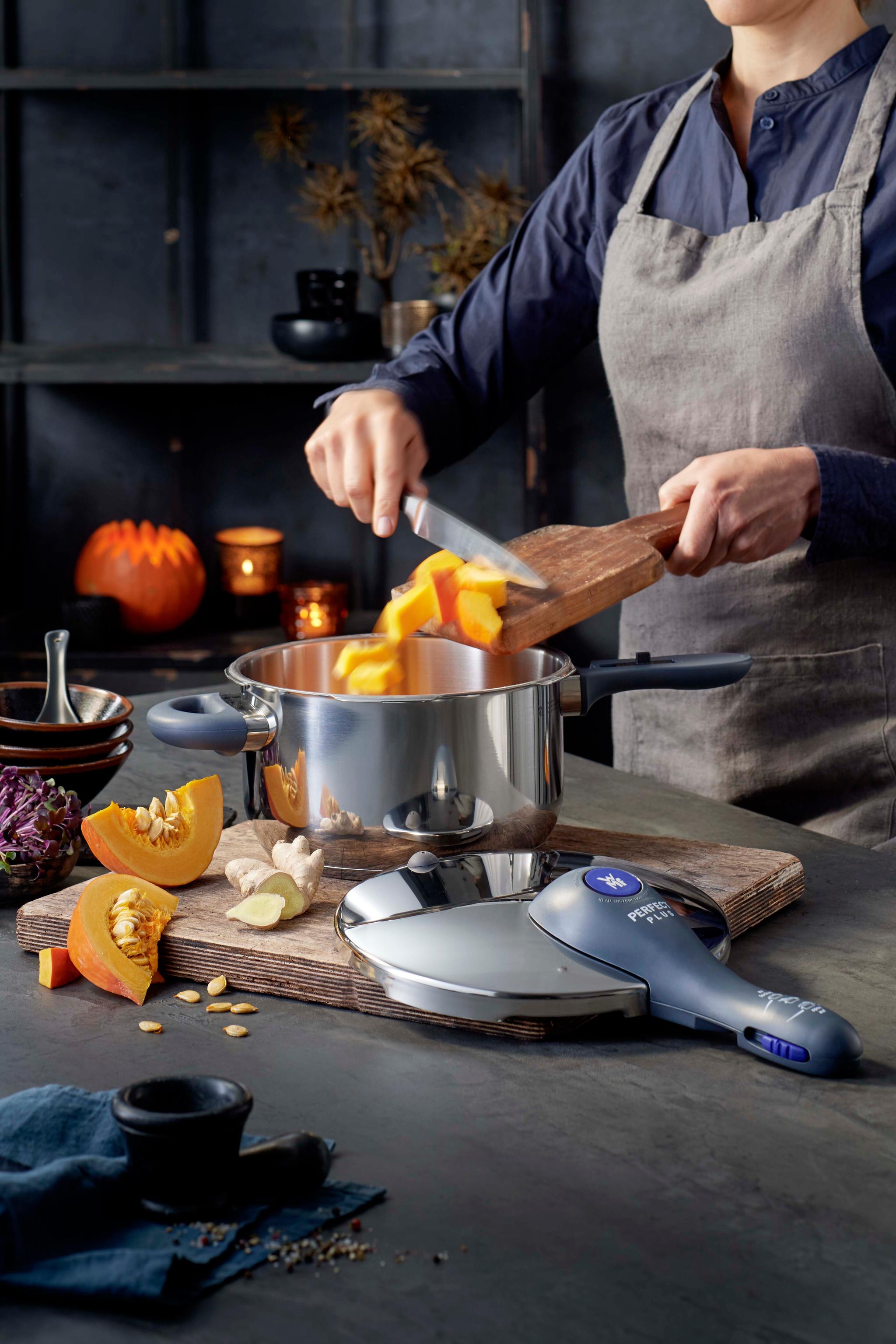 Esiliinapäinen henkilö lisää leikkuulaudalta silputtua kurpitsaa ja inkivääriä ruostumattomasta teräksestä valmistettuun kattilaan, jonka lähellä on painekattilan kansi.